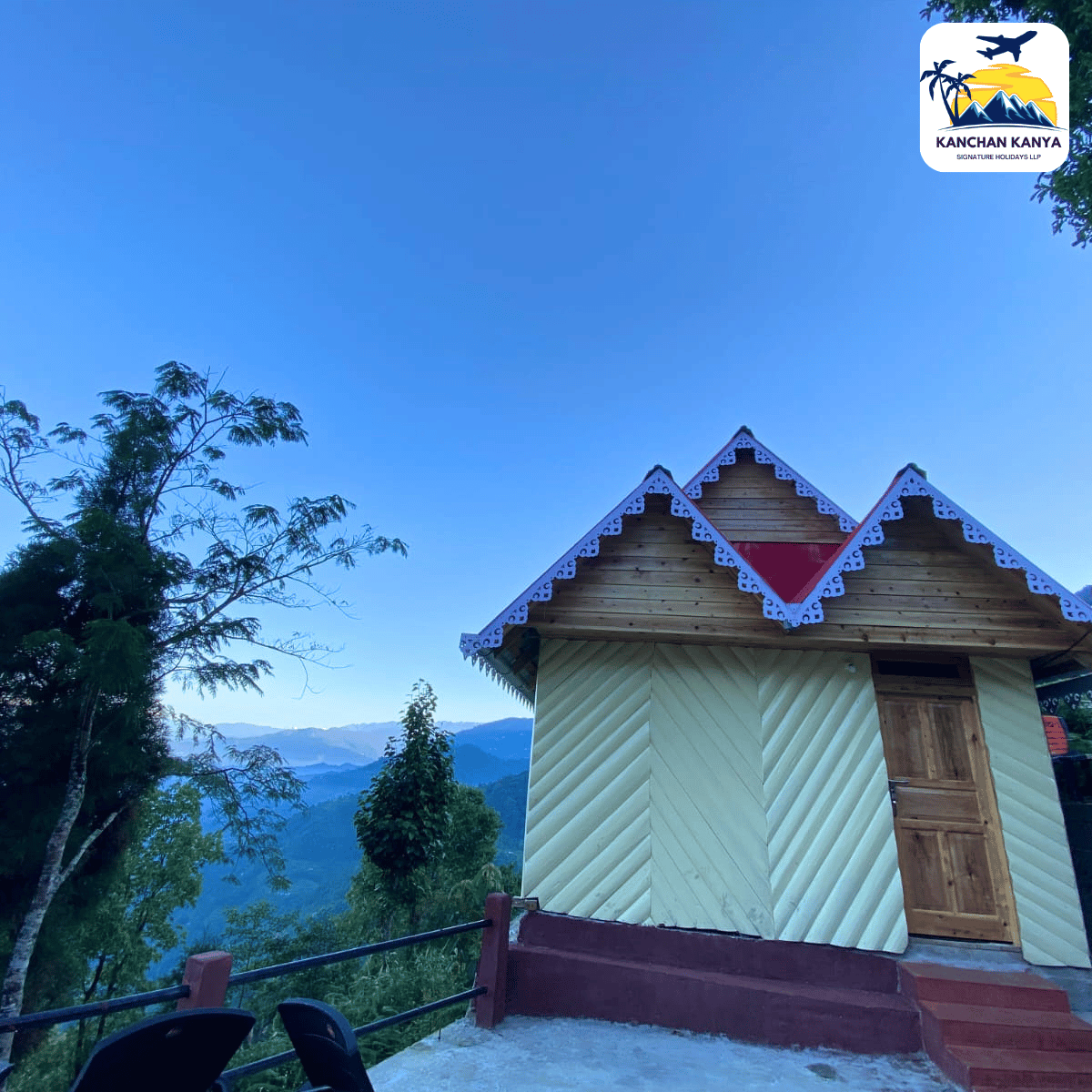 Small wooden cabin with decorative white trim on a hillside, blue sky and distant mountains, travel logo in the top-right corner.