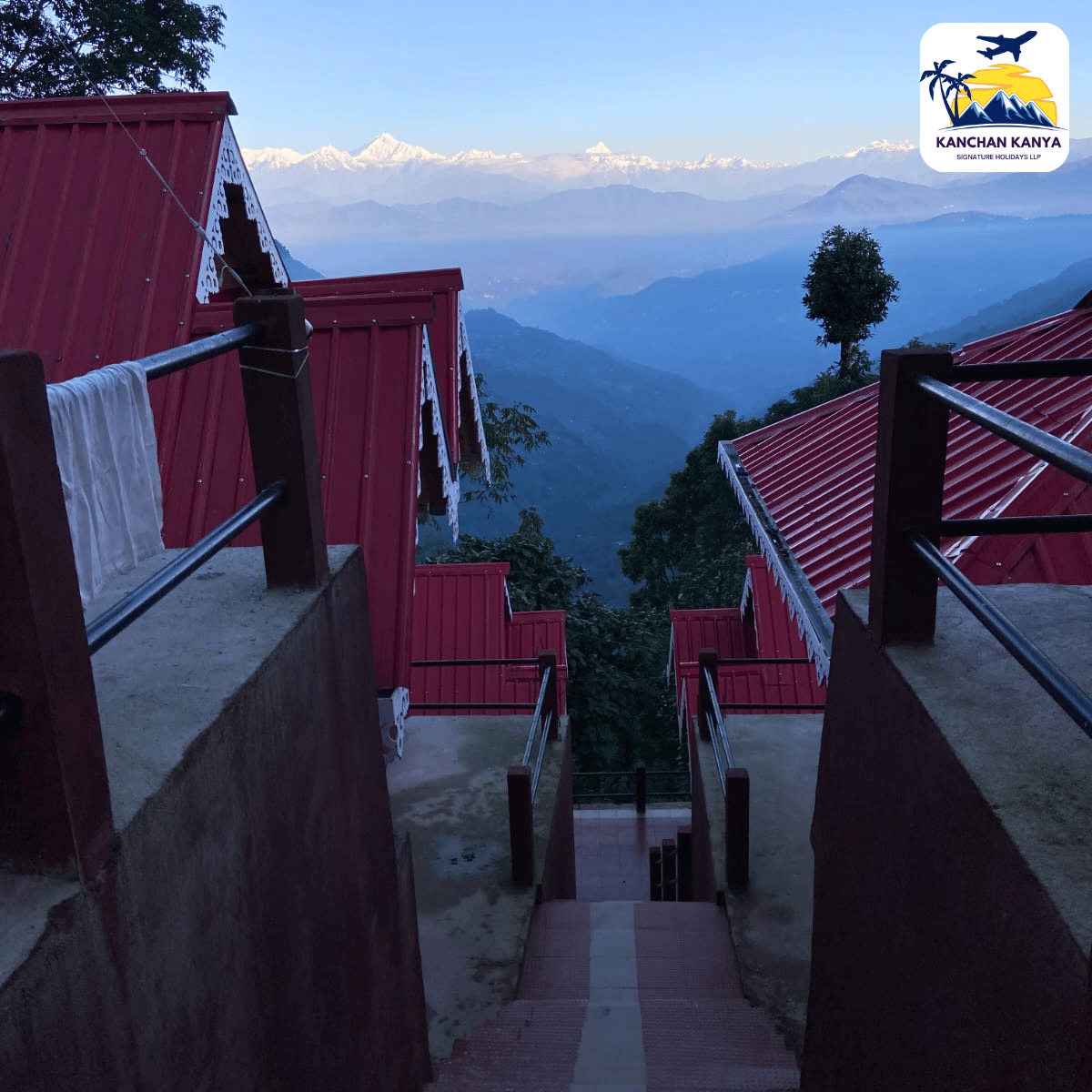 Staircase between red-roofed buildings descends toward a valley with layered blue mountains in the distance; a resort logo sits in the top-right corner.