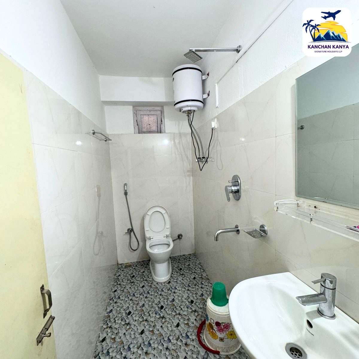 Small bathroom with toilet, wall shower, and water heater mounted on the wall above; pebble floor and white tiled walls, sink visible on the right