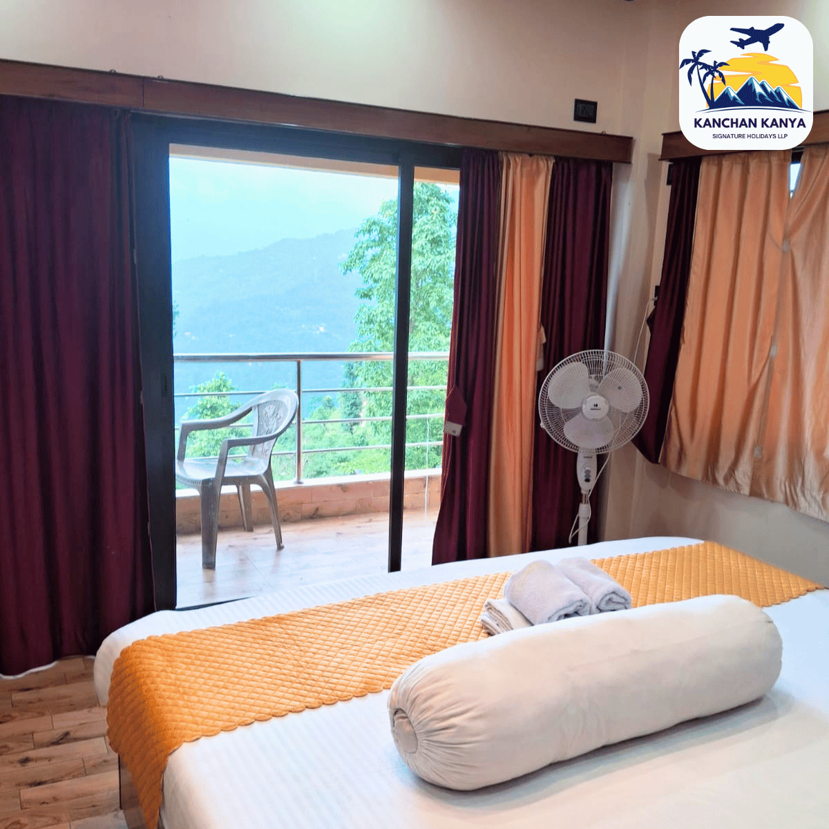Hotel room with balcony door open to a mountain view; bed with orange quilt, rolled towels, standing fan, and maroon/beige curtains on either side, logo in corner.