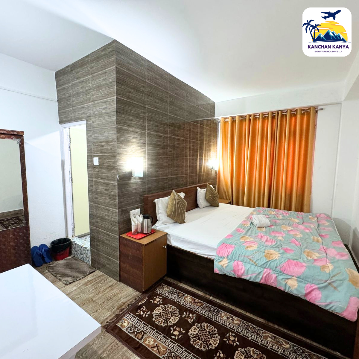 Hotel room with a king bed against a brown wood-panel accent wall, bedside table with a kettle, and orange curtains.
