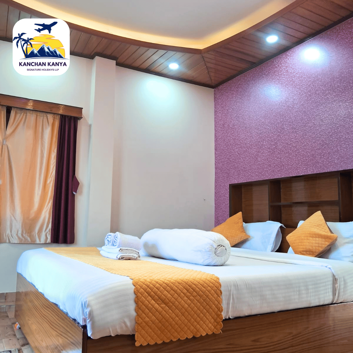 Hotel room with a large bed, orange quilted runner, white linens, and orange pillows; purple textured wall and wooden headboard; window with beige and maroon curtains; ceiling lights; logo in top-left corner