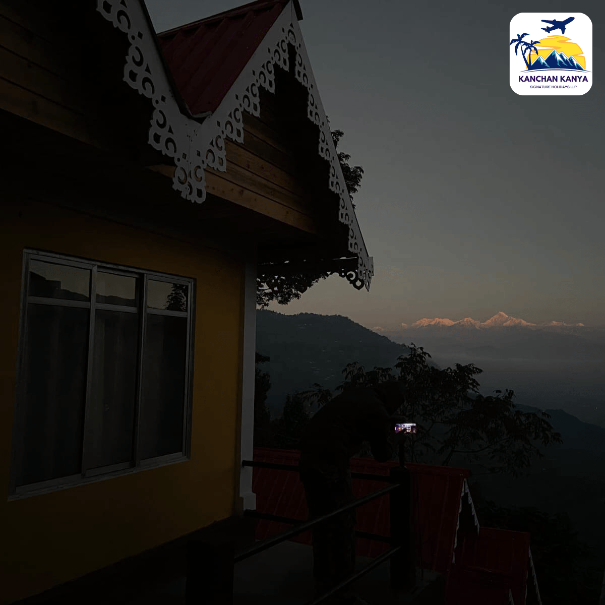 Yellow house with decorative white eave trim at dusk; a person on the balcony takes a photo toward a distant mountain range.