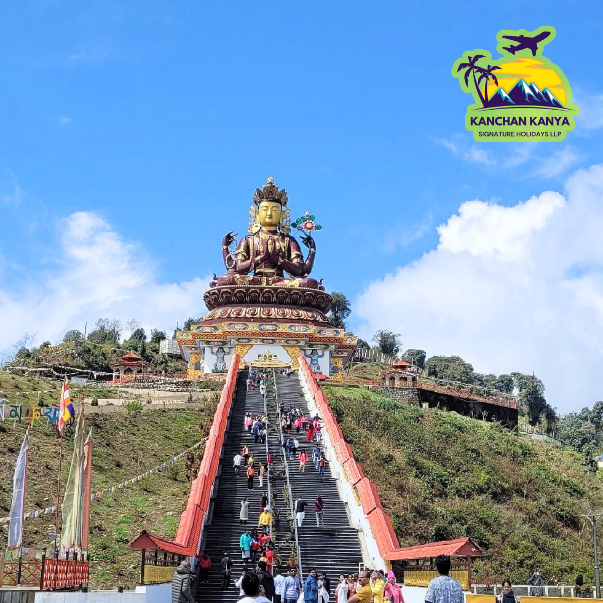 Large seated Buddha statue atop a hill, approached by a long red-bordered staircase filled with visitors, under a blue sky with a travel logo in the top-right corner (KANCHAN KANYA).