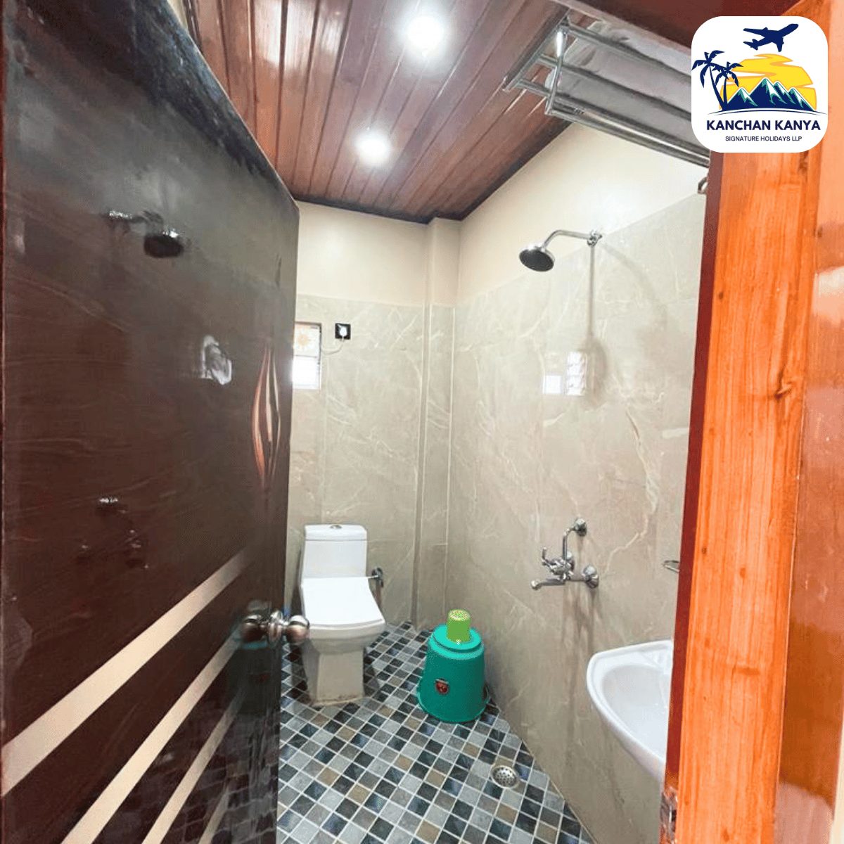Compact bathroom with beige tiled walls, wall-mounted shower, toilet, and sink; green plastic bucket on a blue-gray checkered floor.