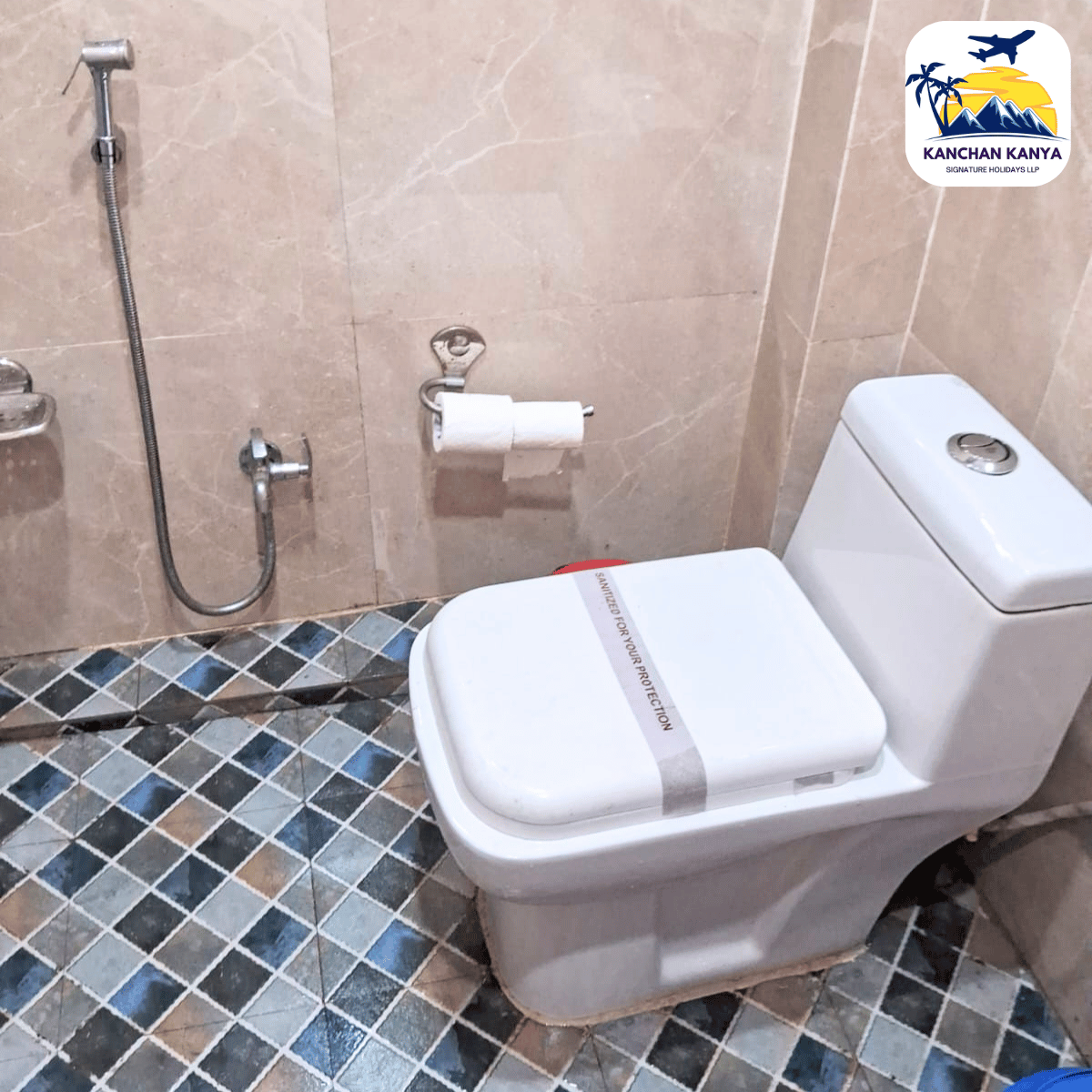 White toilet with a bidet spray, wall-mounted toilet paper holder, and beige wall tiles; blue-gray patterned floor tiles; logo watermark in the top-right corner.
