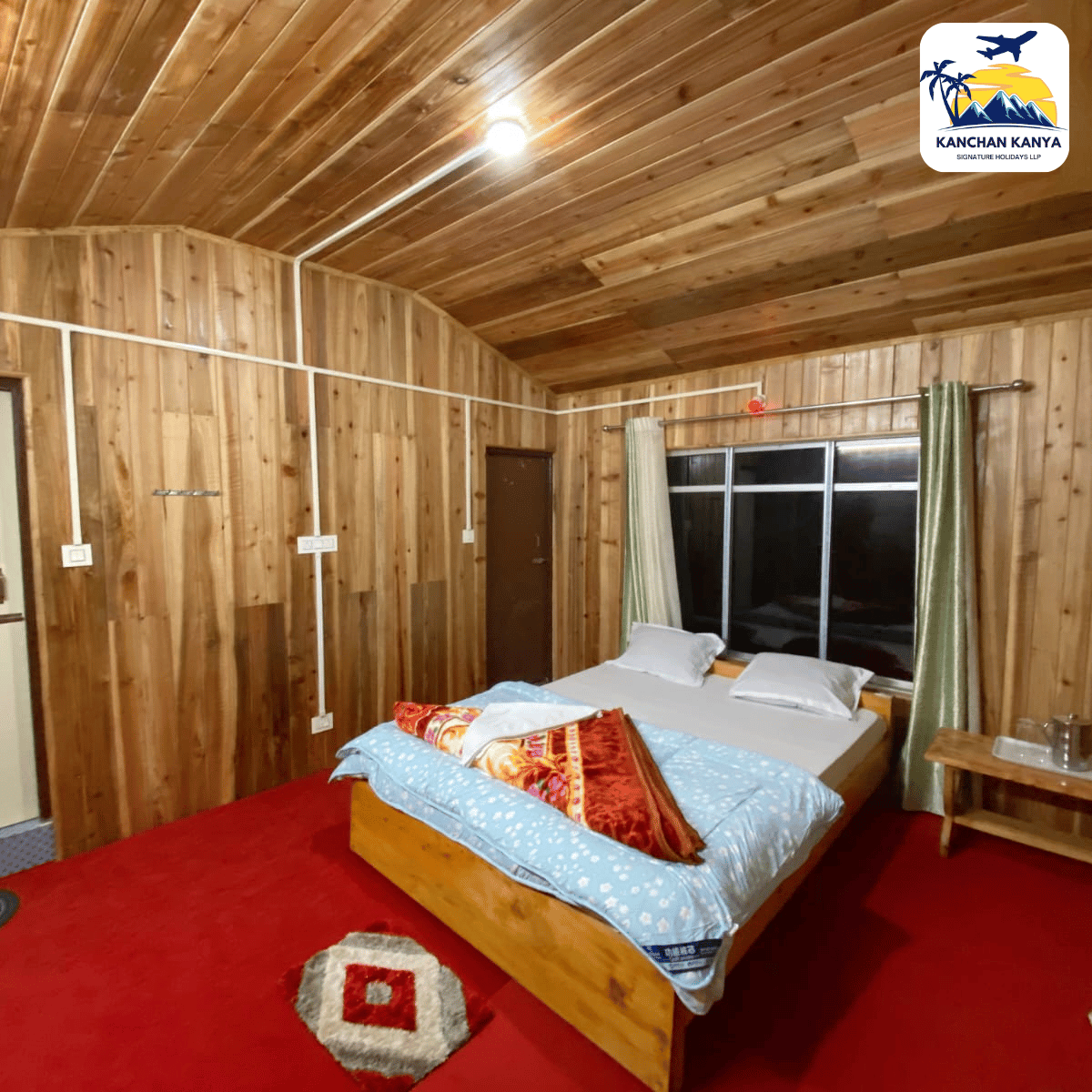 Rustic wooden bedroom with a low bed, blue bedding, red carpet, and a window with green curtains under a curved wooden ceiling. A logo sits in the top-right corner.