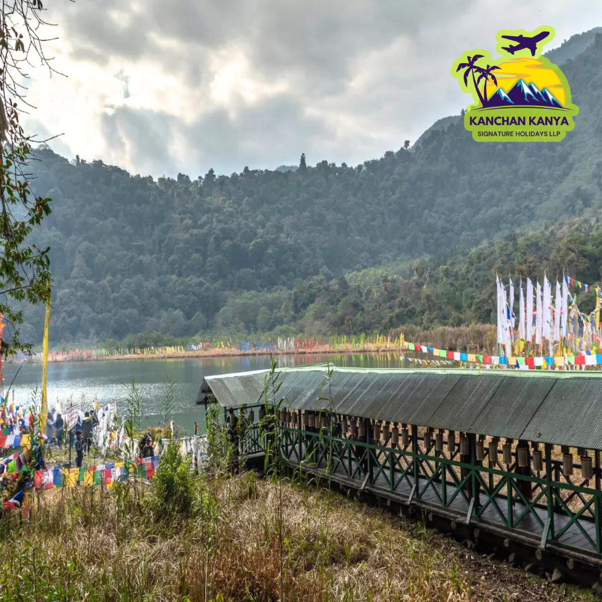 Lakeside scene with a long covered wooden bridge, colorful prayer flags along the shore, and forested hills in the background; travel logo in the top-right corner.