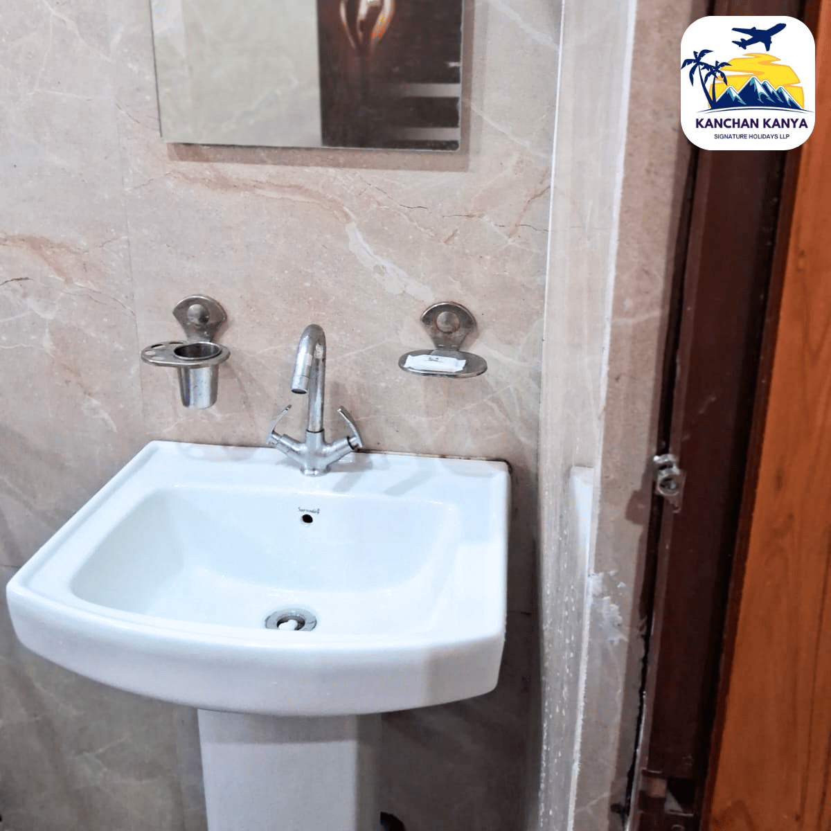 White pedestal sink with a chrome faucet, soap dish and holder on a beige marble-tiled wall; mirror above and a wooden door frame on the right.