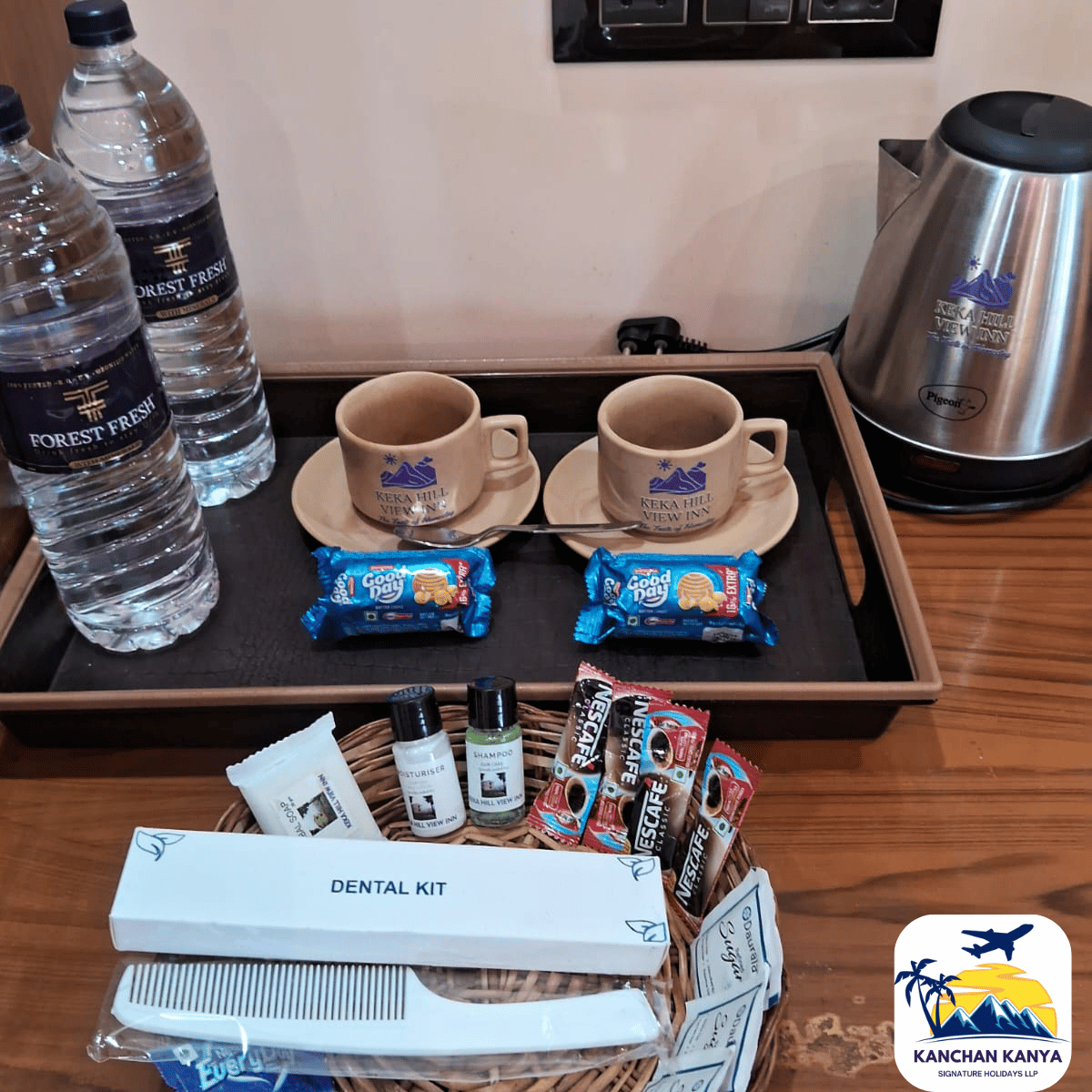 Two beige mugs on saucers with biscuits and water bottles on a wooden tray, plus a metal kettle in the background (hotel tea setup).