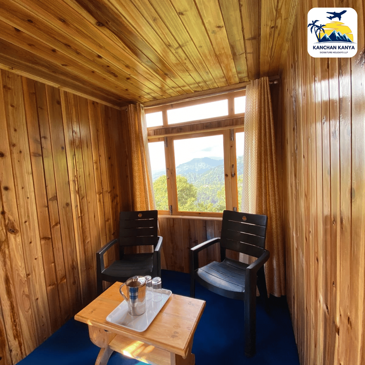 Cozy wood-paneled cabin corner with two black chairs and a small wooden table, looking out to mountains through a window with curtains, logo in the top-right corner.