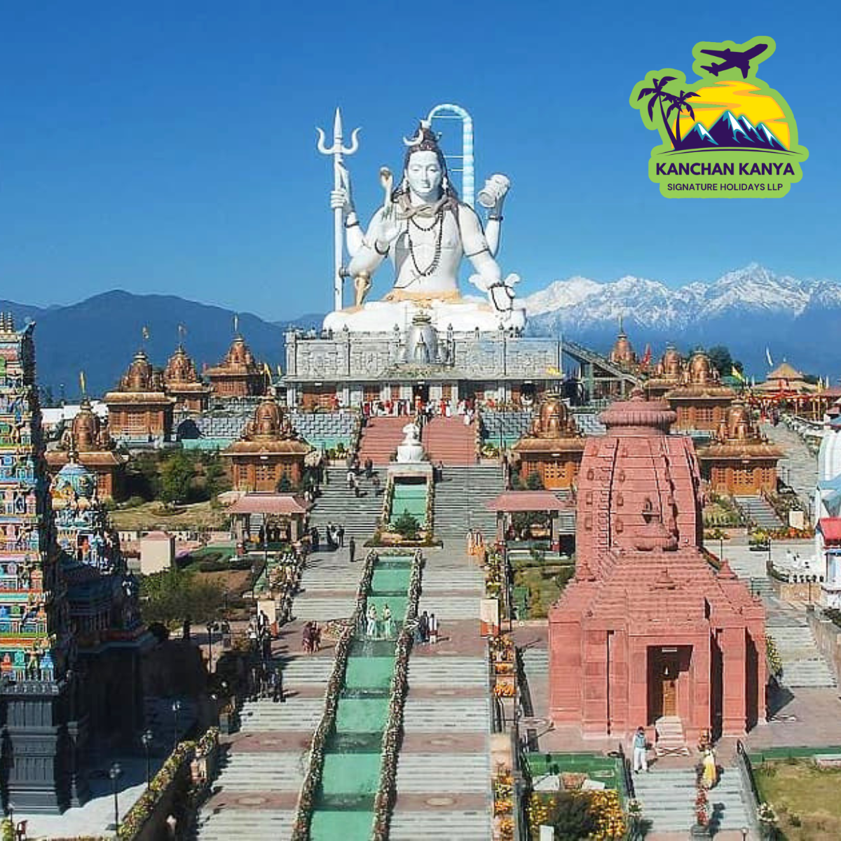 Large Hindu temple complex with a towering white Shiva statue atop steps, flanked by multi-tiered temples and snow-capped mountains in the background; a bright blue sky above.