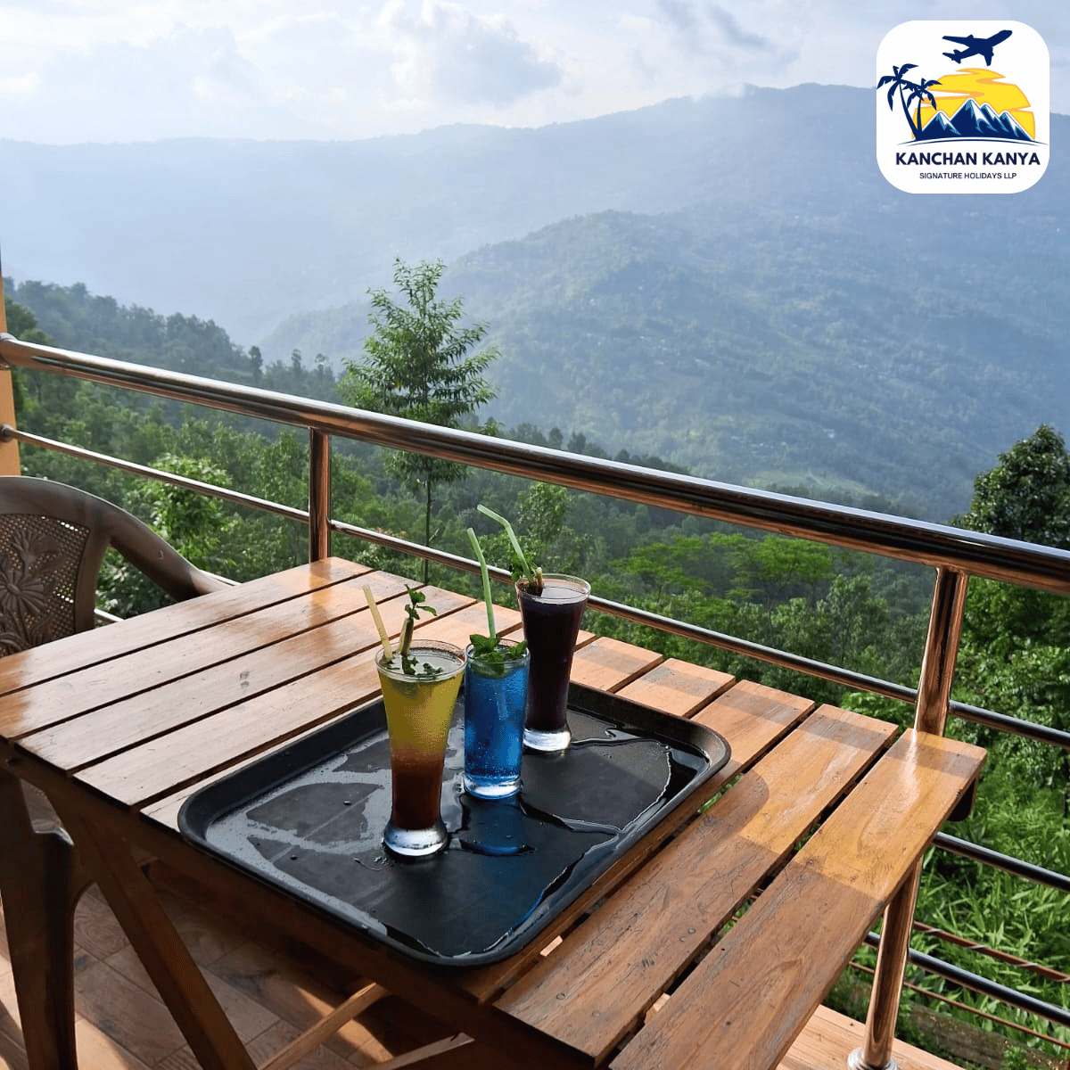 Terrace seating with a wooden table and three colorful cocktails on a tray, overlooking misty green mountains; resort logo in the top-right corner.
