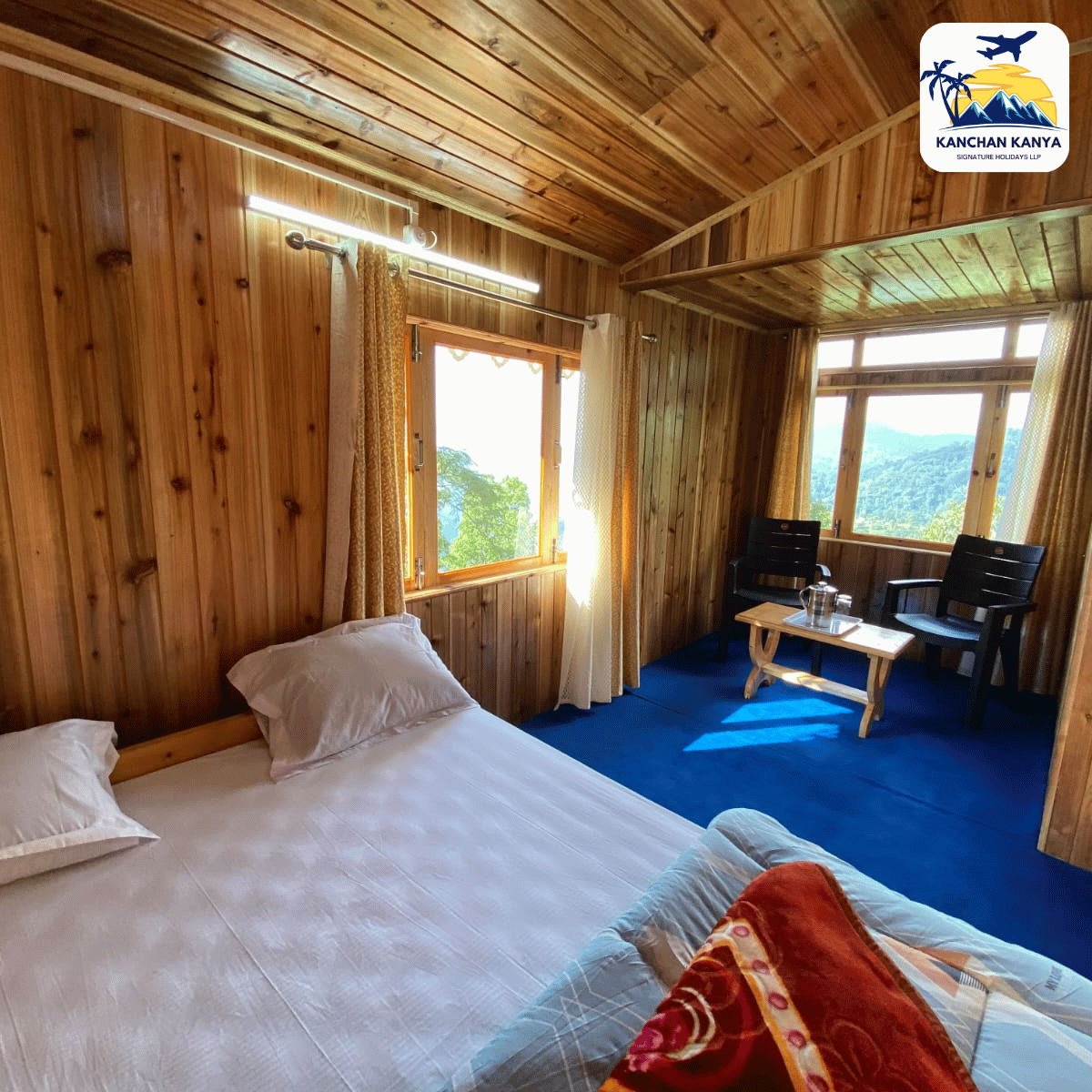 Wood-paneled hotel room with a large bed, blue carpet, and a small seating area by windows overlooking mountains; logo top-right.