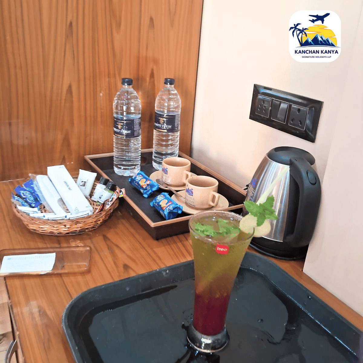 Hotel room refreshment station with two water bottles, snacks, cups, an electric kettle, and a layered mint drink on a tray.
