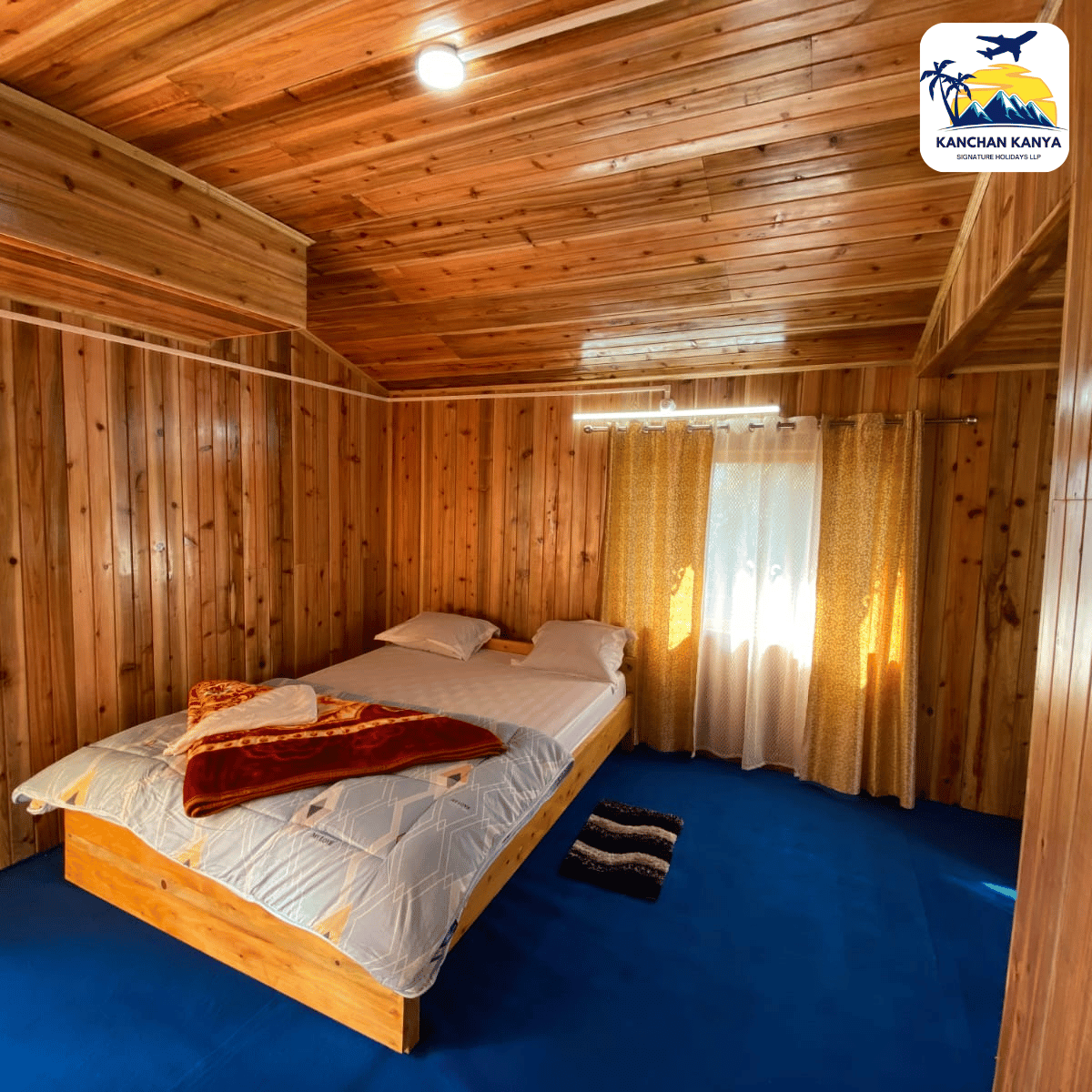 Wood-paneled bedroom with a wooden double bed, white bedding and an orange blanket, set on a blue carpet, next to gold-curtained window. Logo in top-right corner.