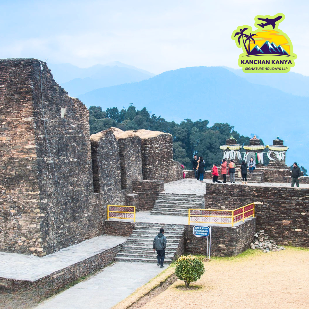 Stone fortress with stairs and a viewing terrace, visitors exploring as blue mountains loom in the background; travel logo in the top-right corner.