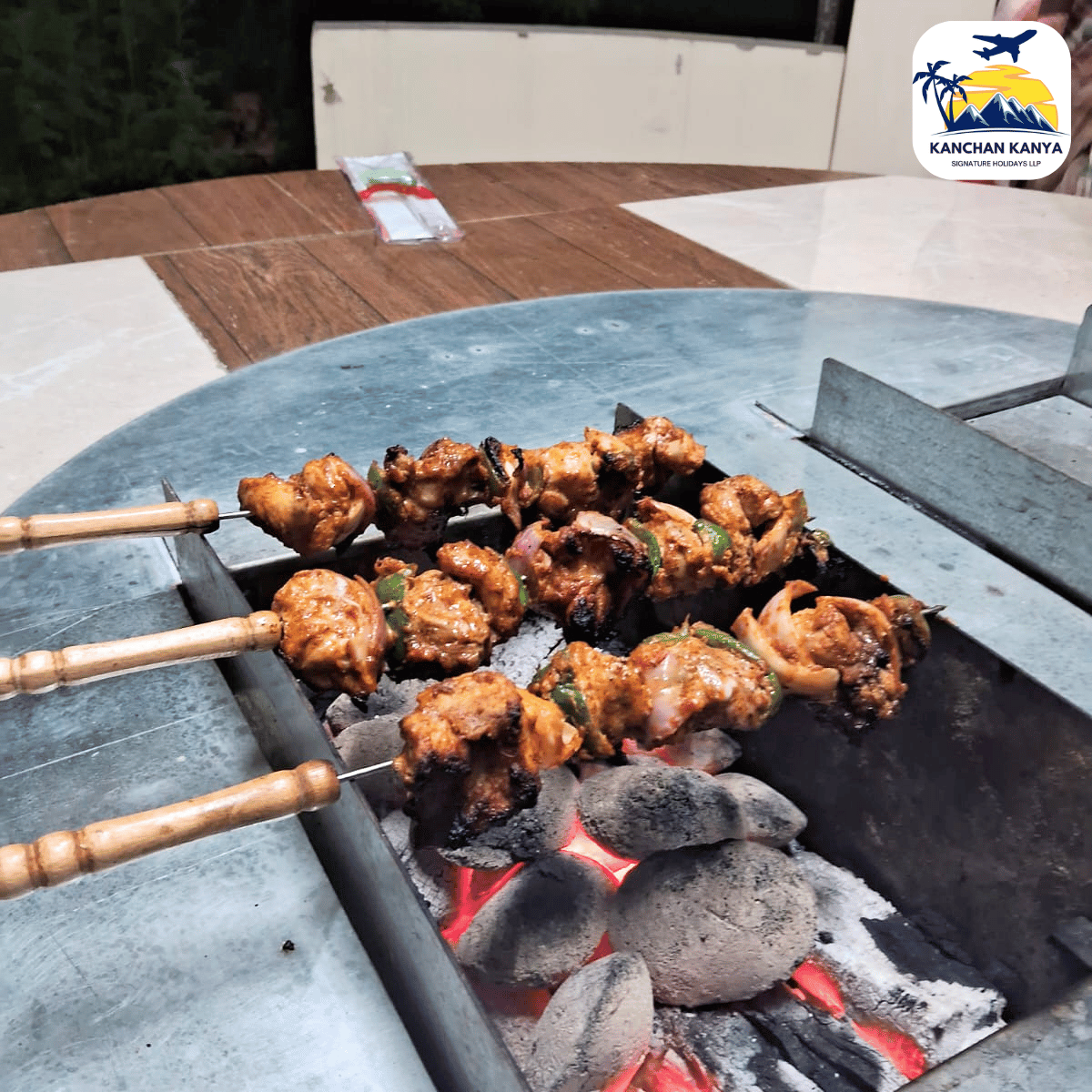 Skewered kebabs grilling over hot charcoal on a round outdoor barbecue; watermark logo in the top-right corner.