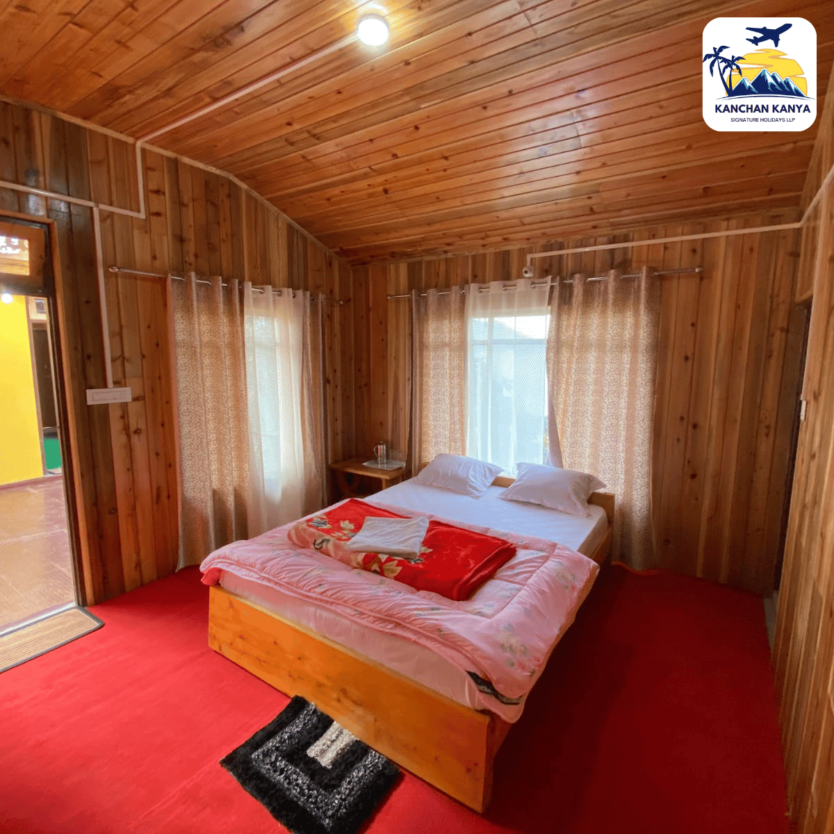 Wood-paneled bedroom with a double bed, white linens and red throw, in a cozy cabin-style room with red carpet and sheer curtained windows.