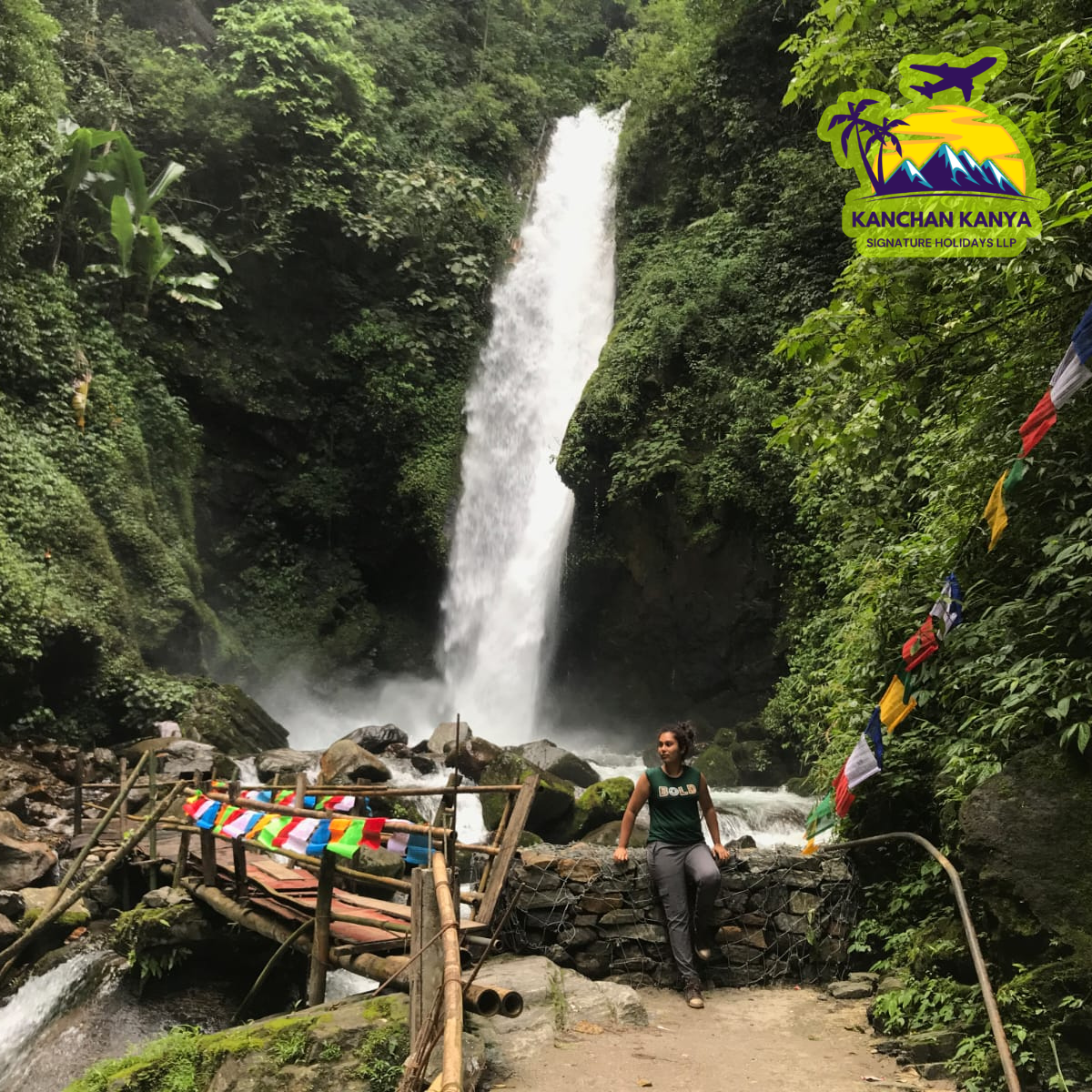 Tall waterfall cascades between green cliffs, with a woman sitting by a stone wall near a colorful flag‑lined path and a wooden bridge nearby.