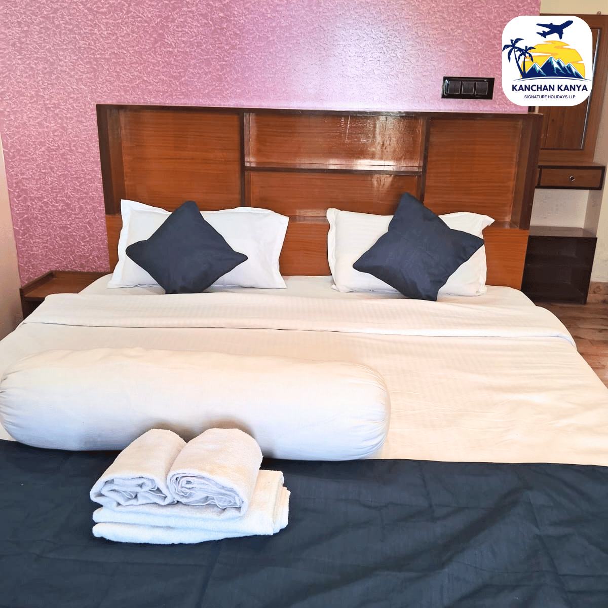Hotel room bed neatly made with white linens, two white pillows and two navy cushions, folded towels at the foot, pink textured wall behind, logo corner.