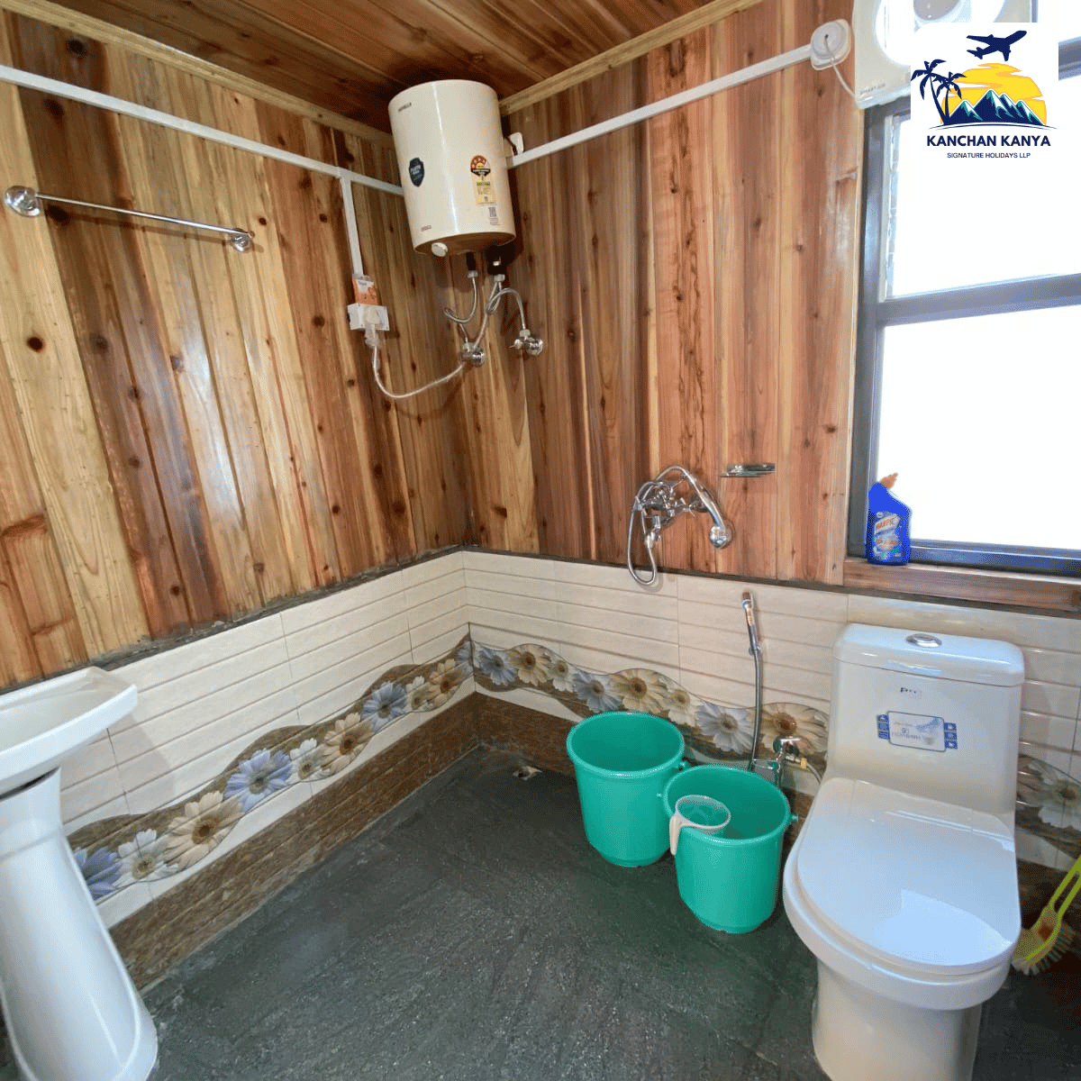 Wood-paneled bathroom with a wall-mounted water heater, sink on the left, and a toilet on the right; two green buckets on the floor.