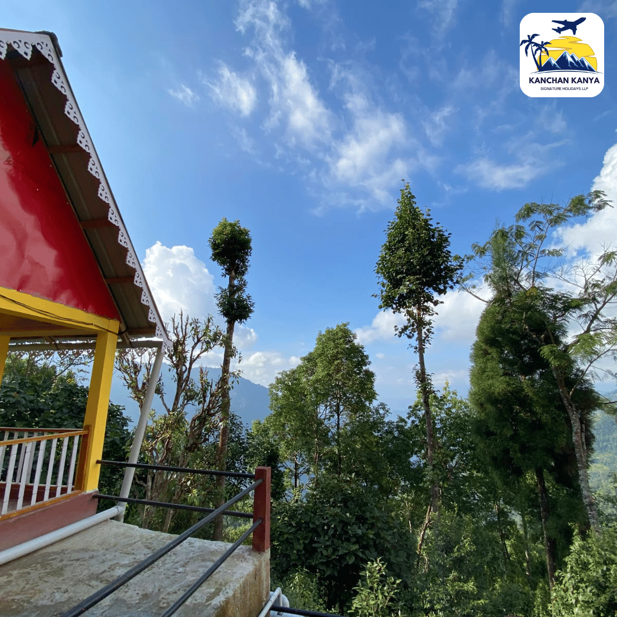 A hillside terrace with a red and yellow house on the left, railing in the foreground, and tall trees against a bright blue sky; logo for Kanchan Kanya in the top-right corner.