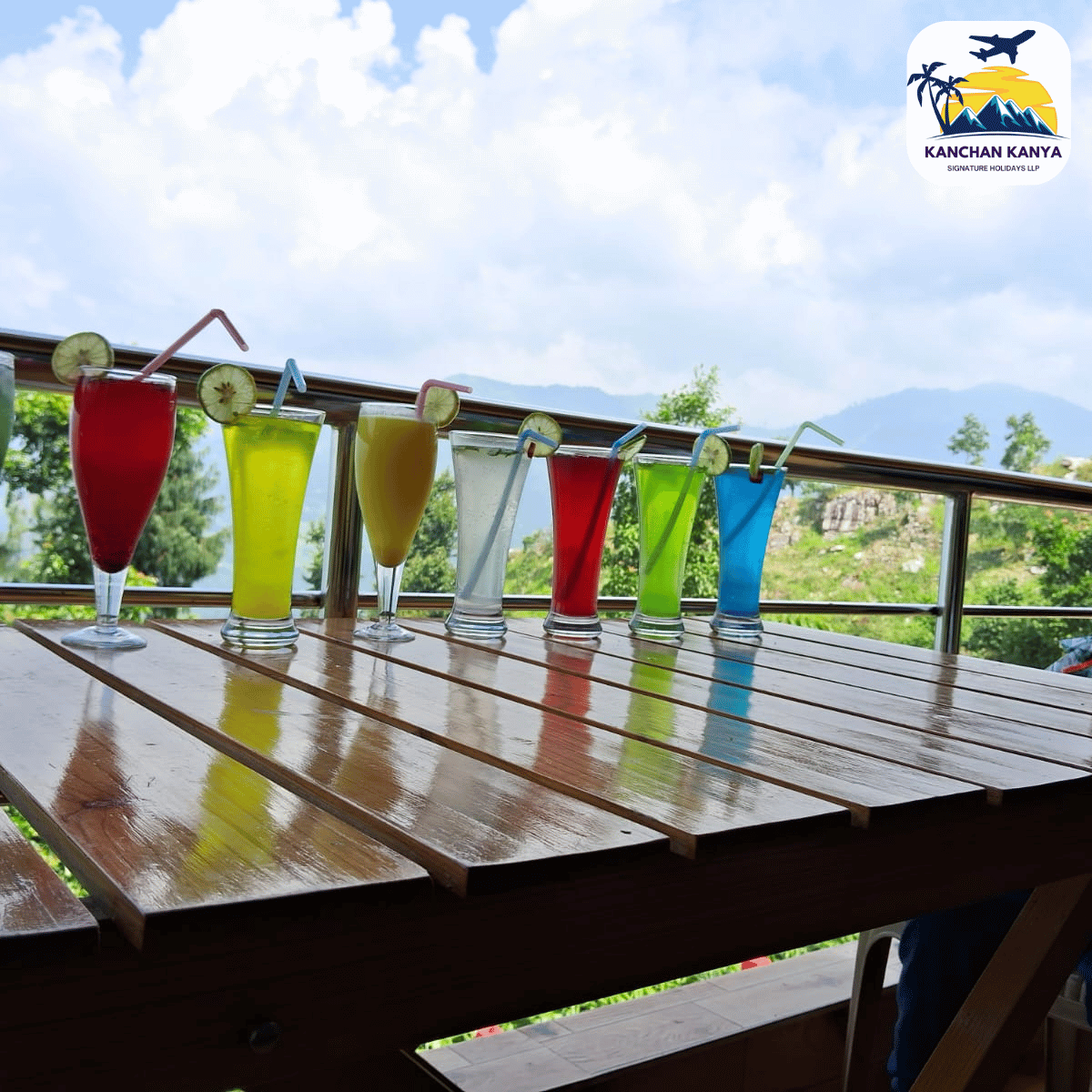 Row of tall colorful fruit drinks on a wooden patio table, with a mountain landscape in the background and a logo in the top-right corner.