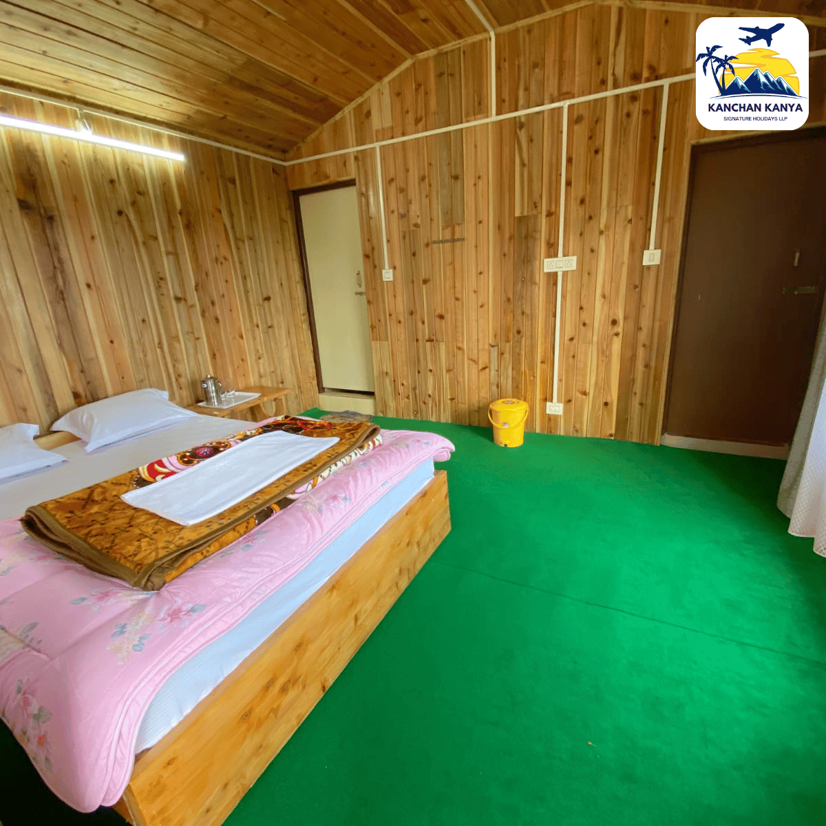 Wood-paneled hotel room with a double bed, pink bedding, a small bedside table and kettle, green carpet, and a door at the far wall; a yellow bucket near the wall.