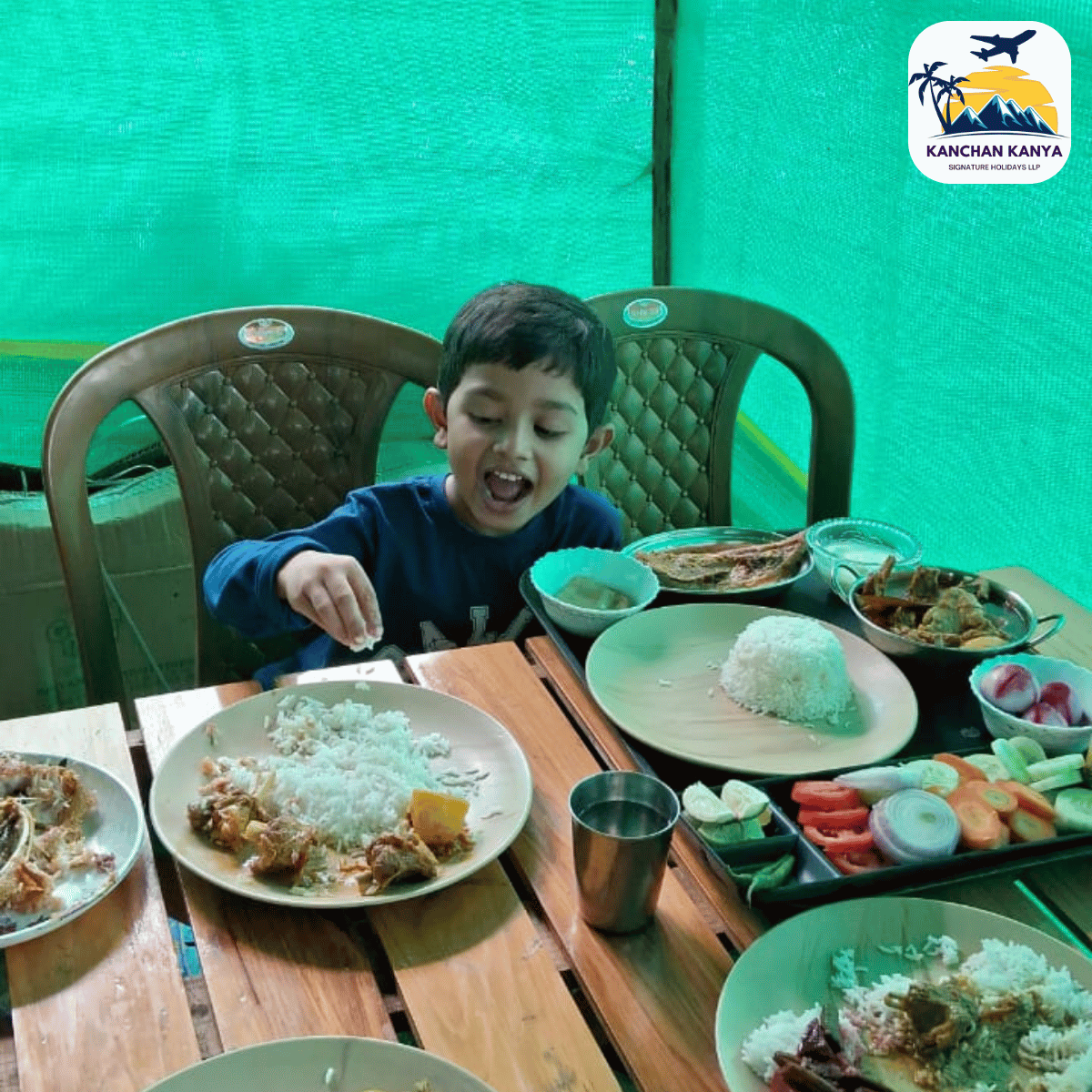 Young boy sits at a wooden table, smiling as he eats a meal with rice and curries, surrounded by several plates of food. There is a teal-green backdrop and a logo in the top-right corner.