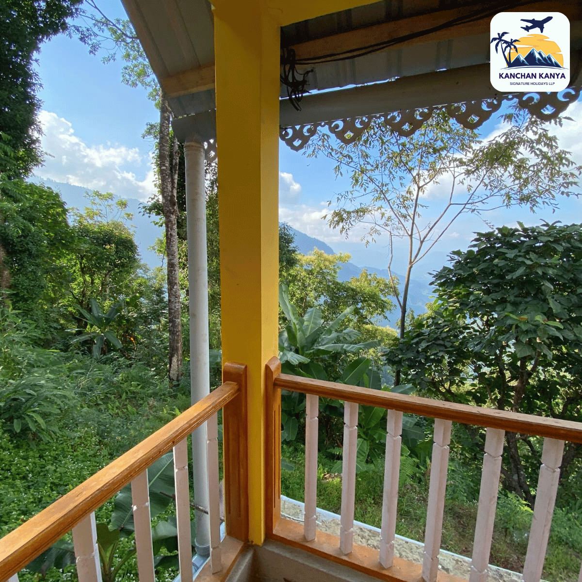 Balcony with yellow posts and wooden railing overlooks a lush tropical garden and distant mountains; logo appears in the top-right corner.