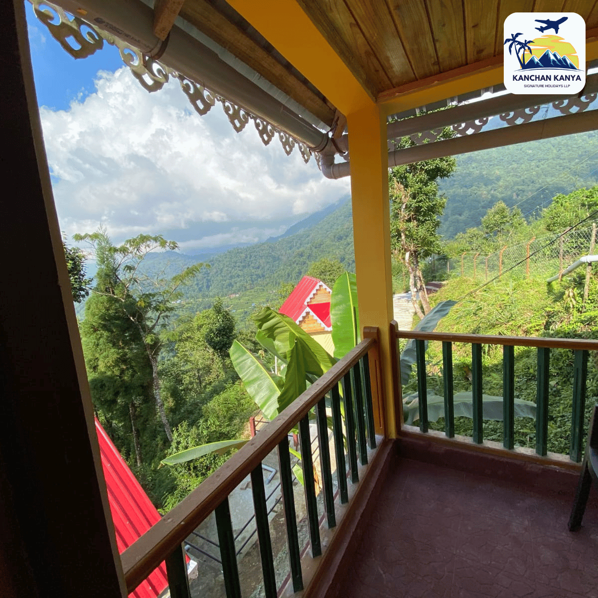 Terrace/balcony with a wooden railing overlooking green hills and distant mountains; yellow support column in view. Logo visible in the top-right corner.