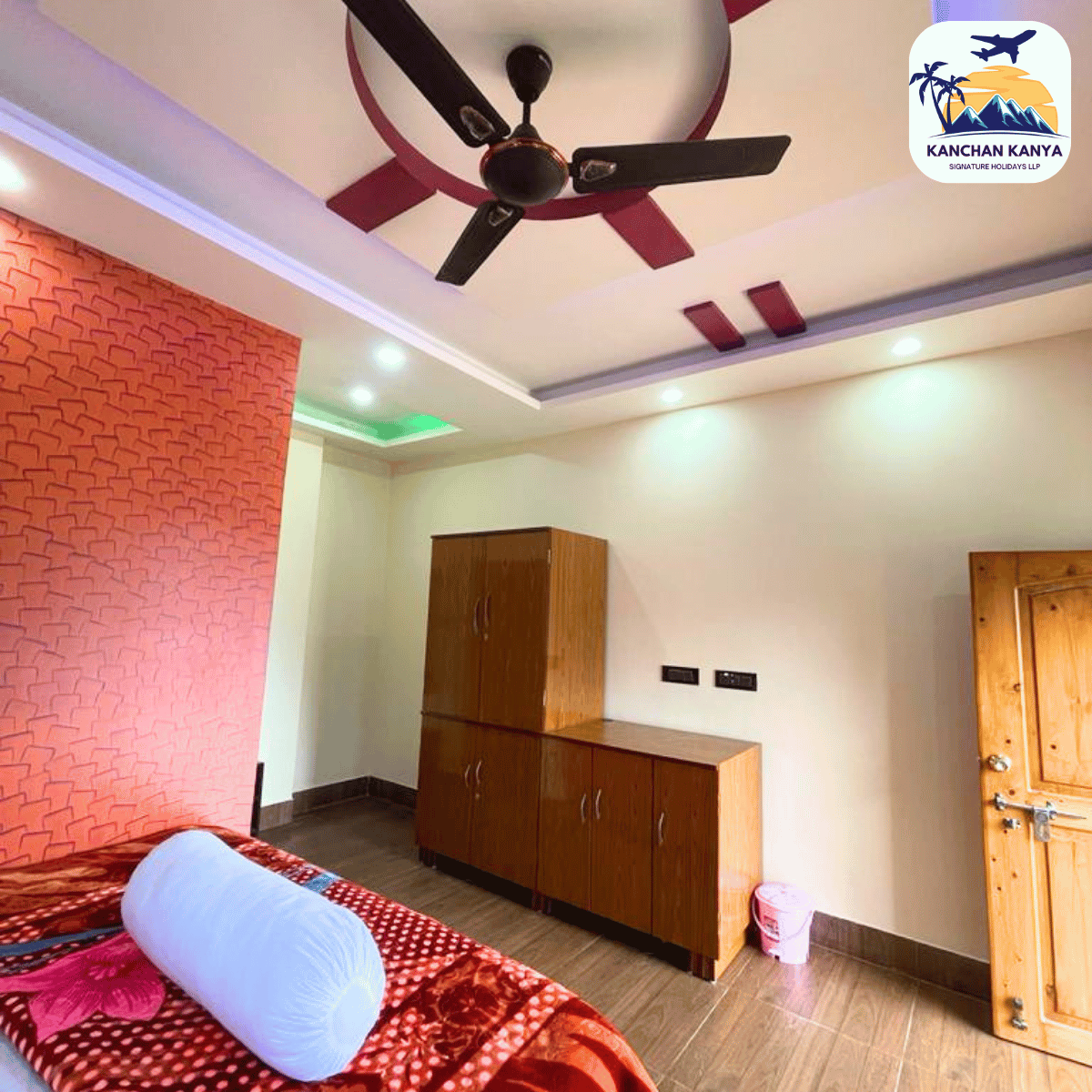 Bedroom with ceiling fan, pink textured wall, and wooden cabinet set; bed with red patterned bedding and a light blue bolster pillow in the foreground.