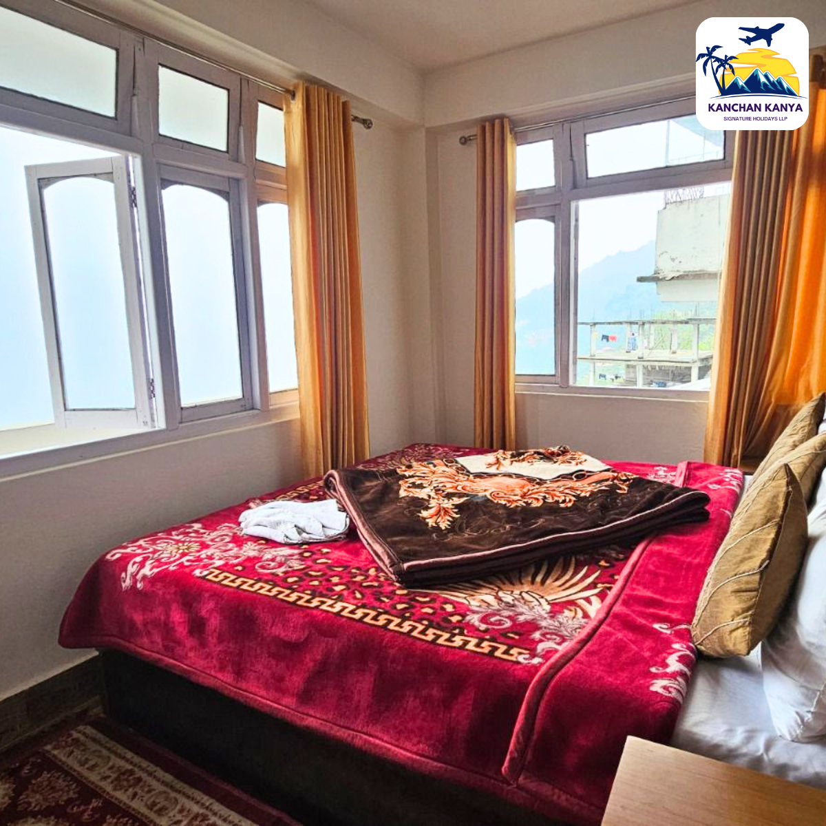 Hotel bedroom with a red patterned bedspread, folded brown blanket, and cream pillows beside large windows with orange curtains.