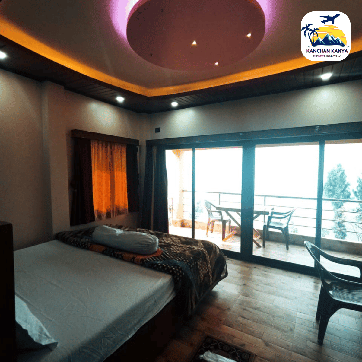 Hotel bedroom with a king bed, patterned quilt, and a window with orange curtains facing a balcony door.