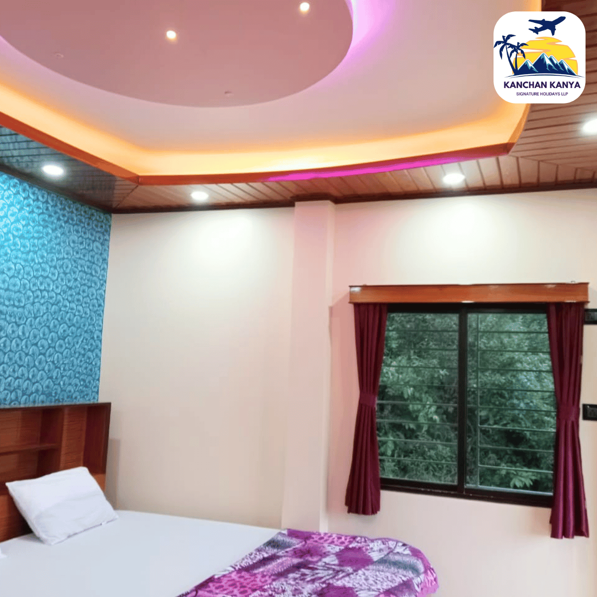 Hotel room interior with a white bed, pink walls, blue patterned accent wall, burgundy curtains, and a round lit ceiling; Kanchan Kanya logo top-right.