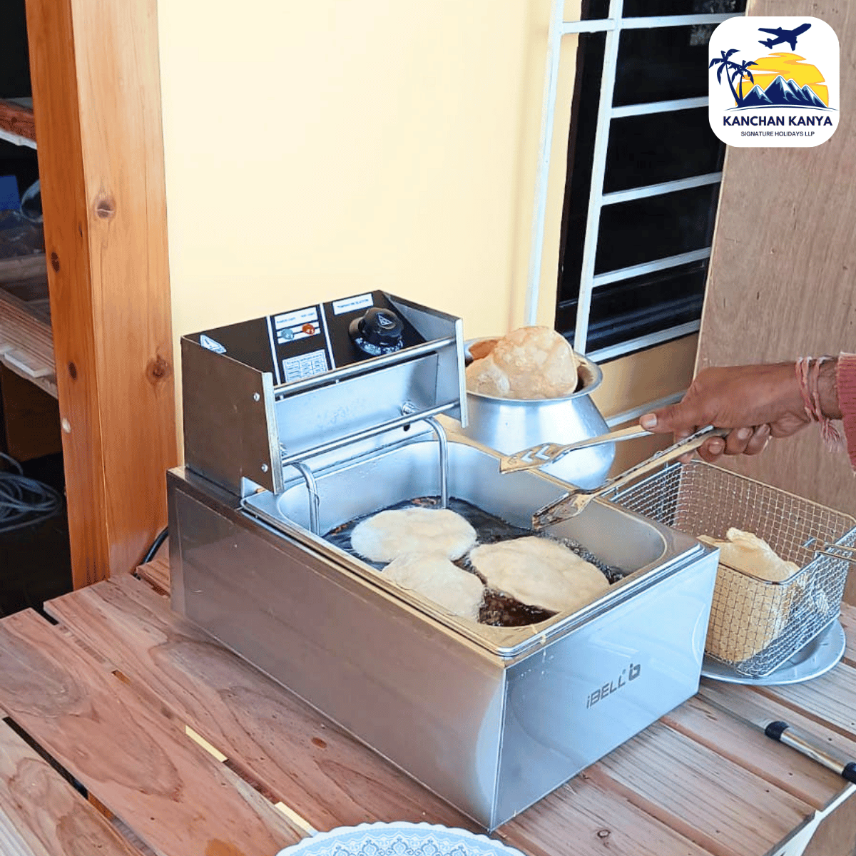 Commercial deep fryer on a wooden table frying round dough pieces; a person uses tongs to lift a fried item, with a logo in the corner.