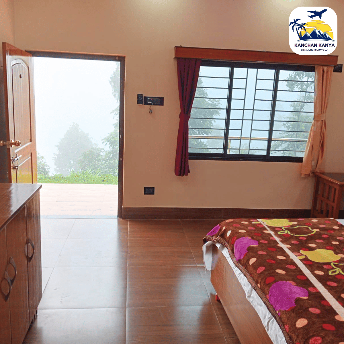 Hotel room interior with a wooden dresser, single bed with a brown patterned blanket, and a large window with red and beige curtains; door open to a foggy outdoor view of trees.