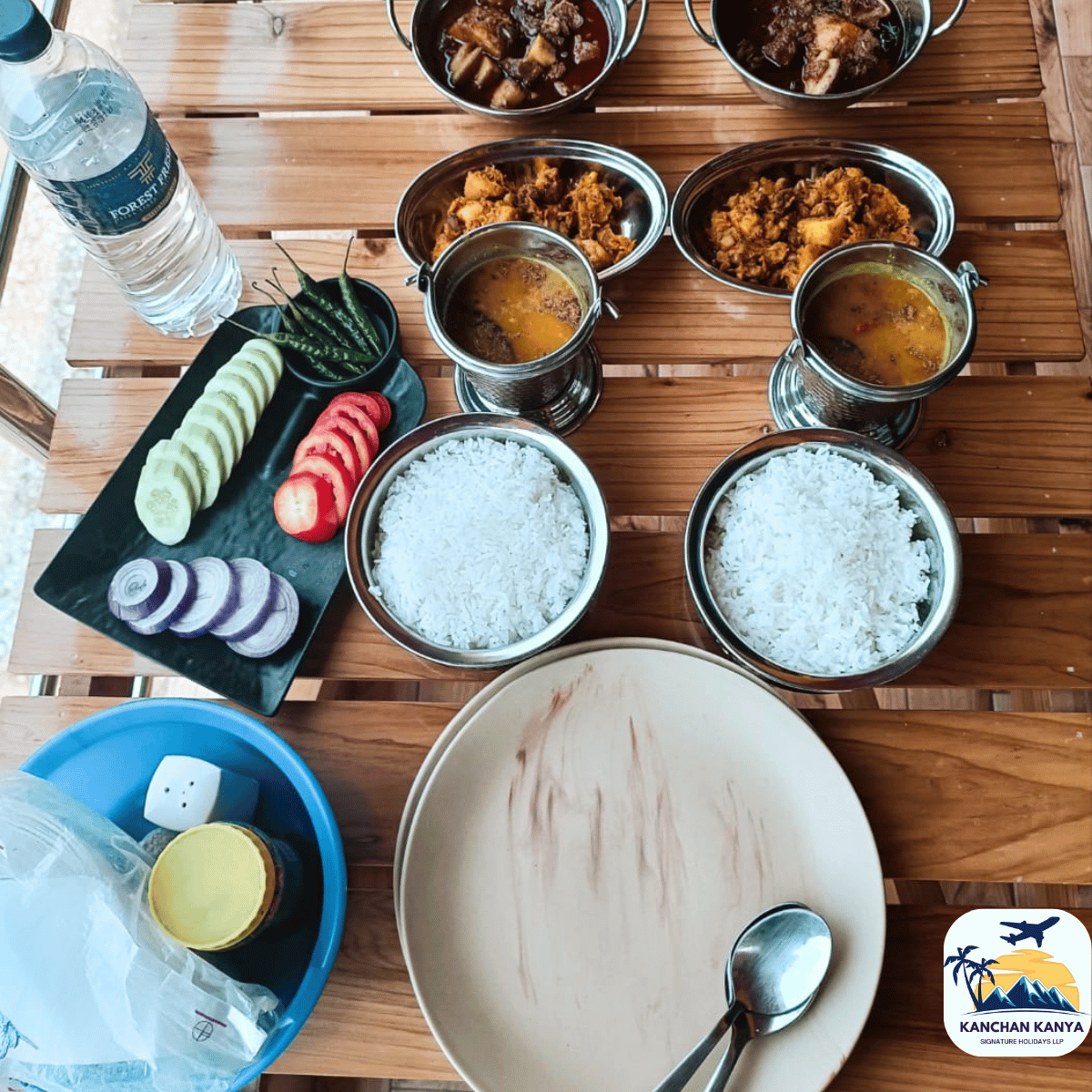 Communal Indian meal spread on a wooden table: metal bowls of curry and rice, sliced vegetables on a slate platter, water bottle nearby, and a logo in the corner.