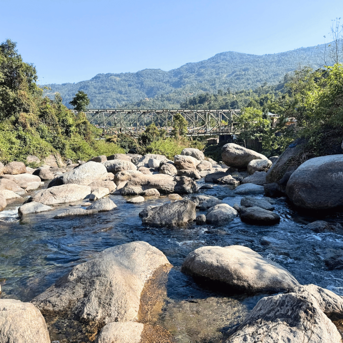 Rocks and a flowing river in the foreground with a truss bridge and forested hills in the background.