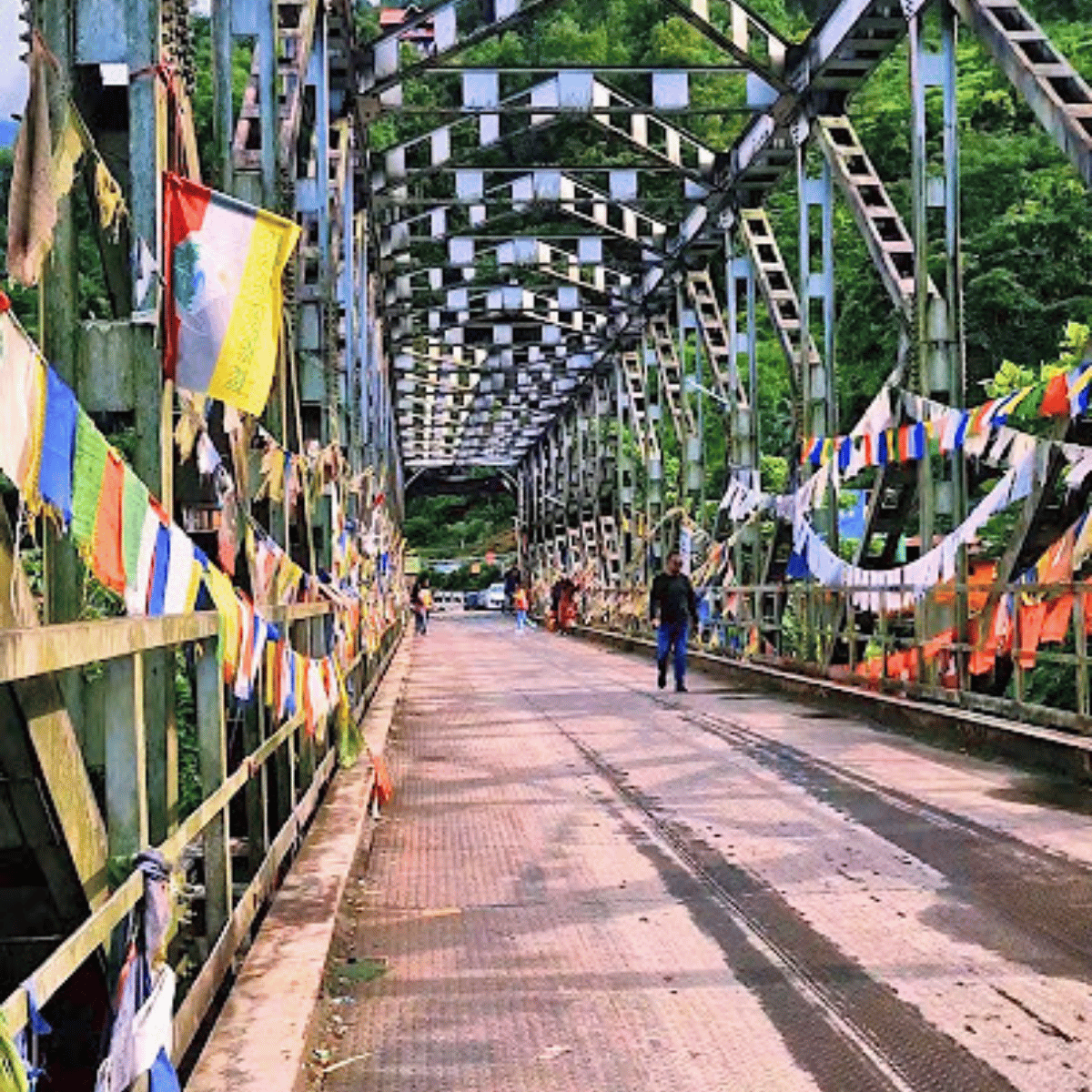Pedestrian walking across a long metal truss bridge decorated with colorful prayer flags and banners, surrounded by greenery.