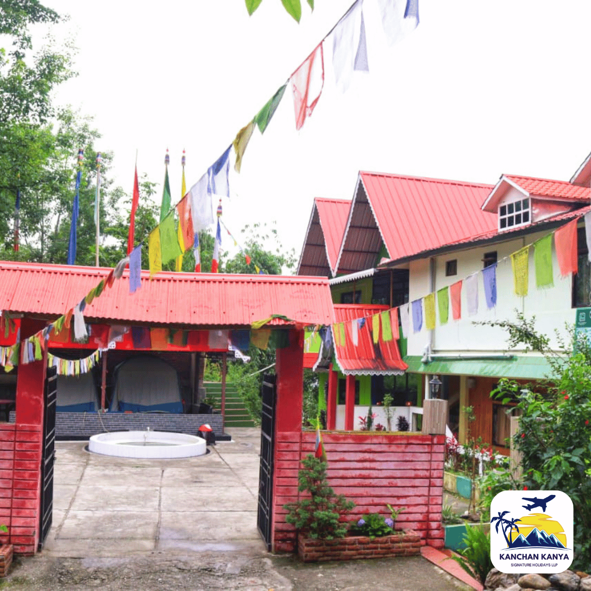 Colorful resort with red-roofed buildings, string of colorful flags, and a circular fountain in the courtyard; logo in bottom-right corner.