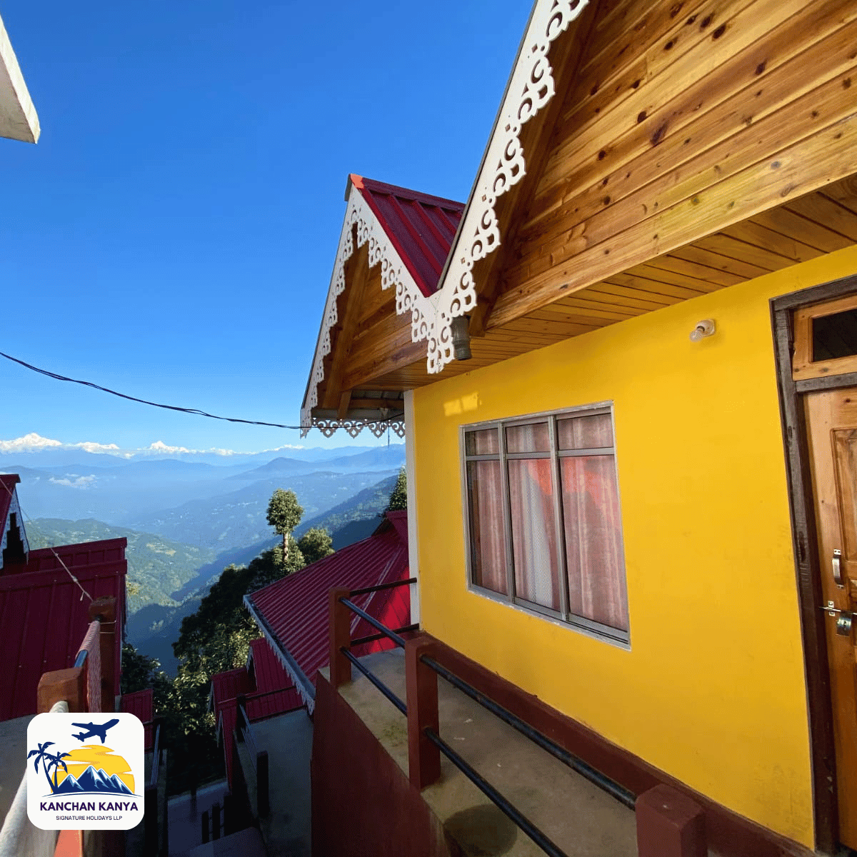 Yellow hillside house with ornate white trim on a wooden gable, railing in foreground, mountains and red-roofed buildings in the valley behind.