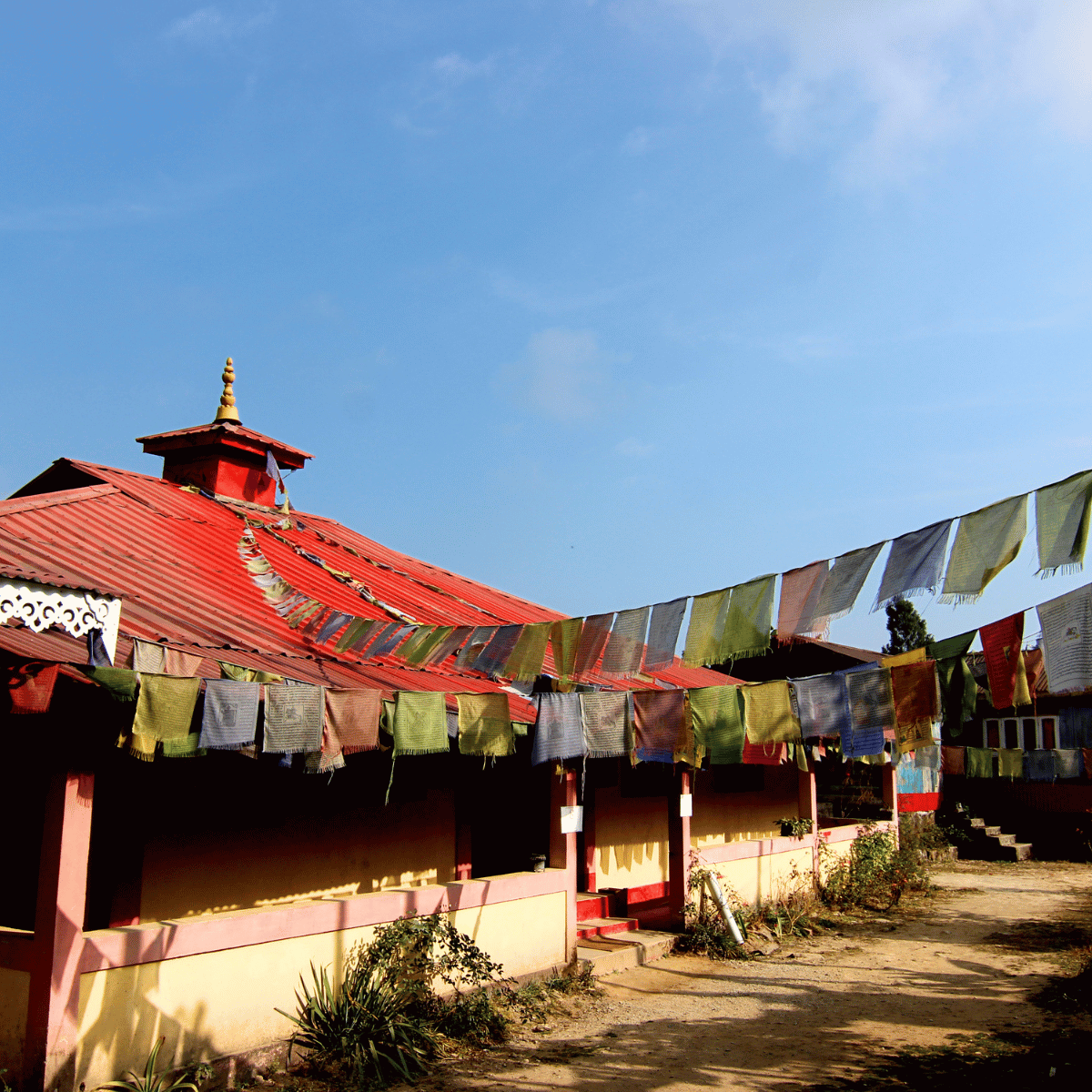 Temple exterior with a red roof and colorful prayer flags strung across a sunny courtyard against a blue sky.