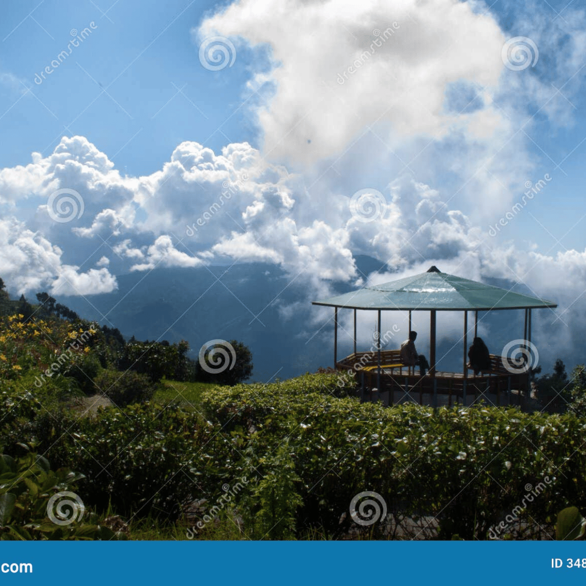 Open-air gazebo on a grassy hillside with two people seated, overlooking a dramatic cloud-filled sky and distant mountains.