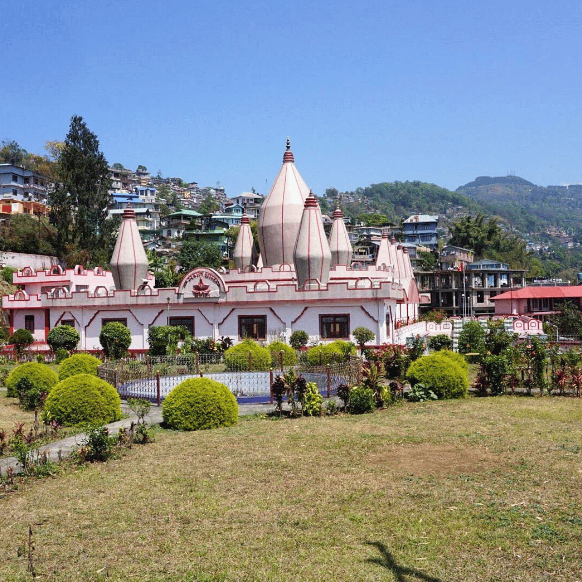 Pink and white temple with multiple dome towers set in a garden, hillside houses in the background under a clear blue sky.