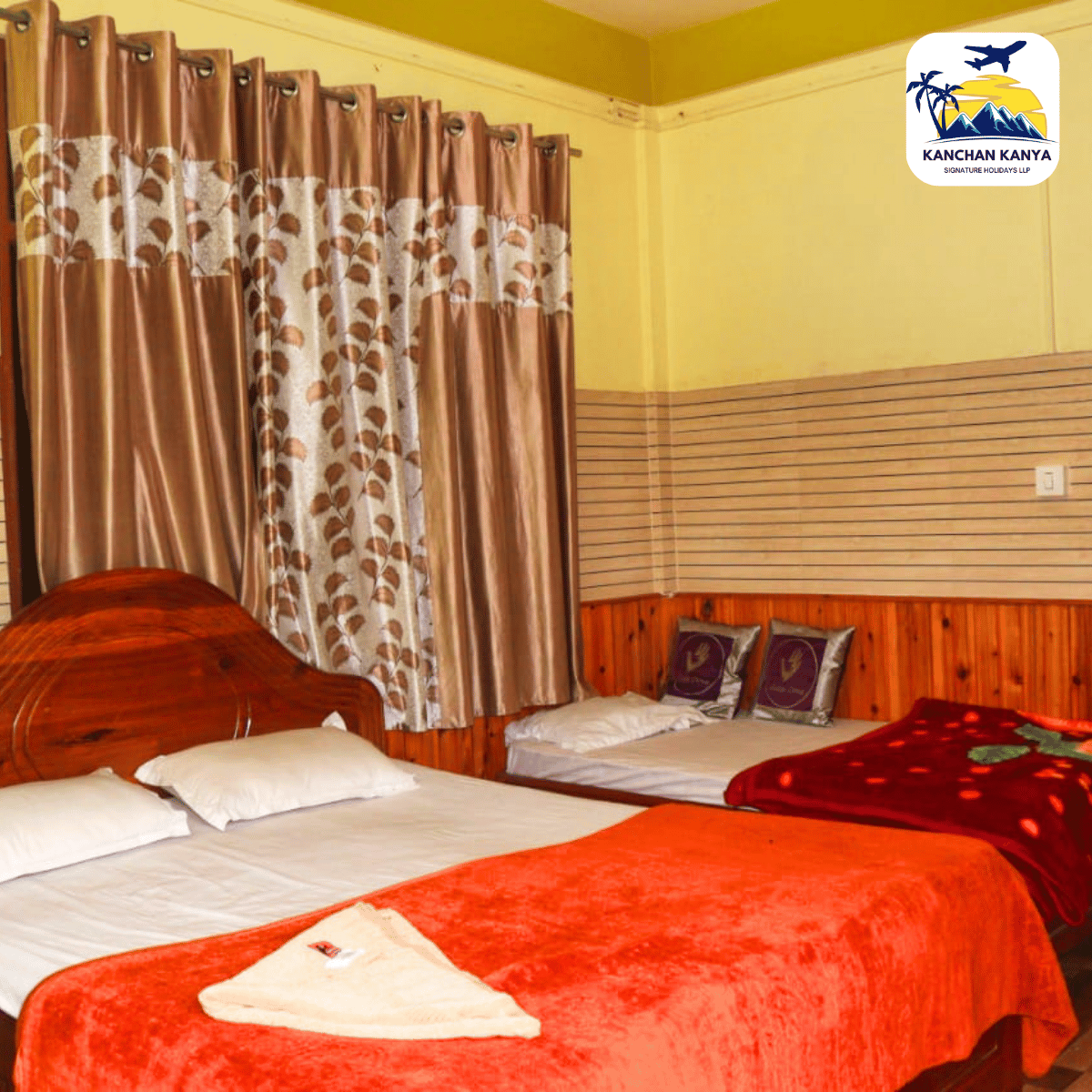 Hotel room with two twin beds, white pillows, red bedspreads, and a towel folded at the foot of one bed. Wood-paneled wall and patterned curtains; logo in top-right corner.