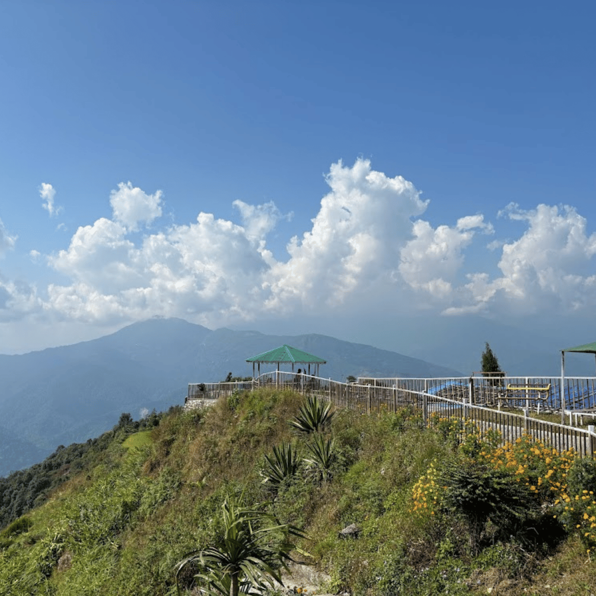 Hilly overlook with a green-roofed gazebo, metal railing, and flowering shrubs, facing distant mountains under a blue sky with clouds