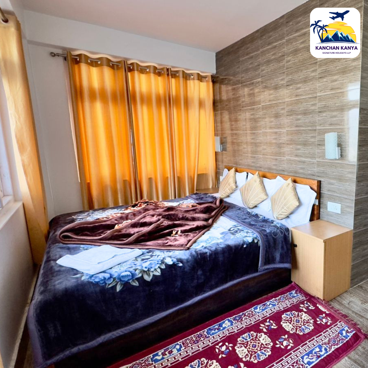 Hotel-style bedroom with a wooden bed, white pillows, beige decorative cushions, amber curtain panels, and a red patterned rug; brand logo visible in the top-right corner.