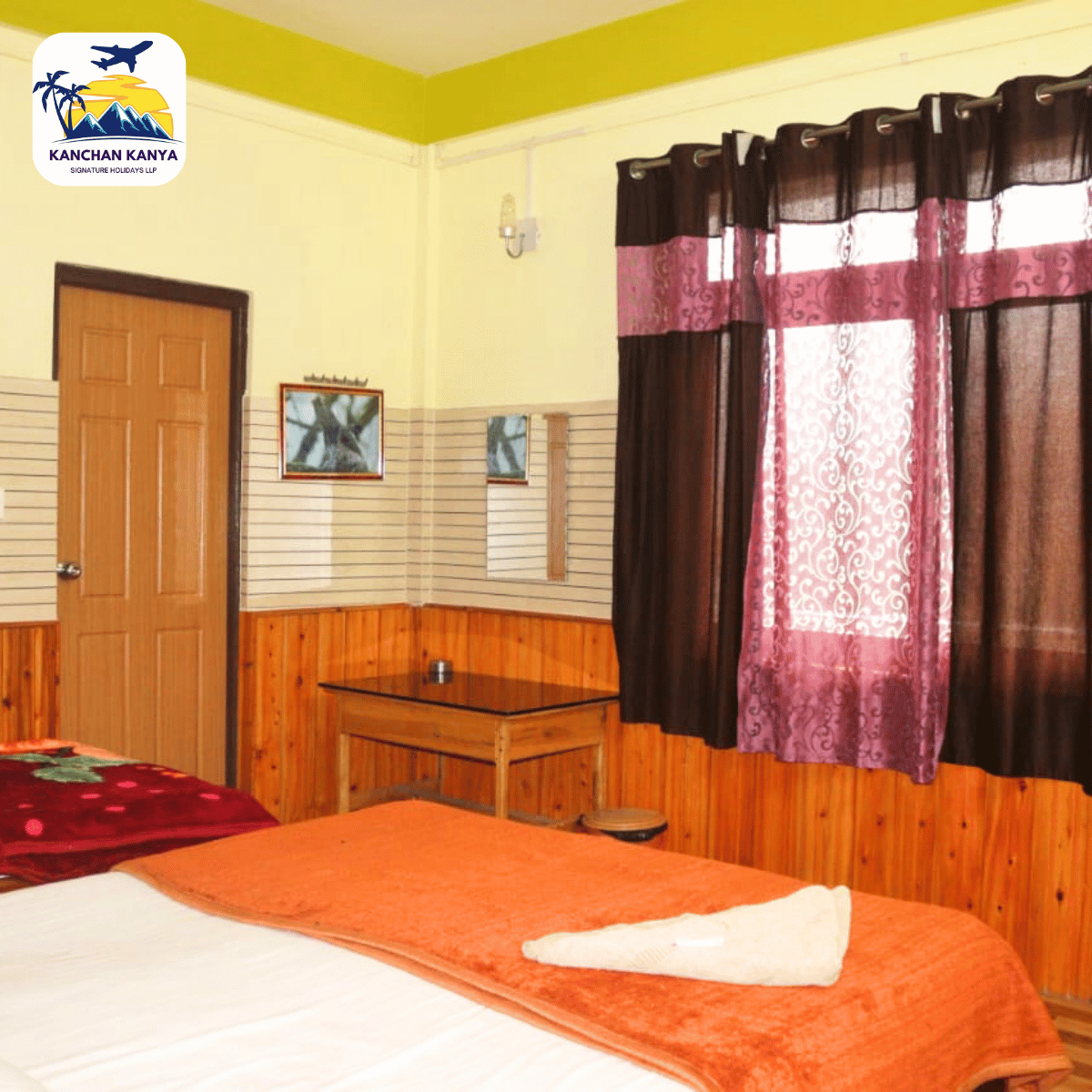 Hotel room interior with orange bedspreads, wood-paneled walls, a small desk, wall mirror, and purple curtains; logo in the top-left corner.